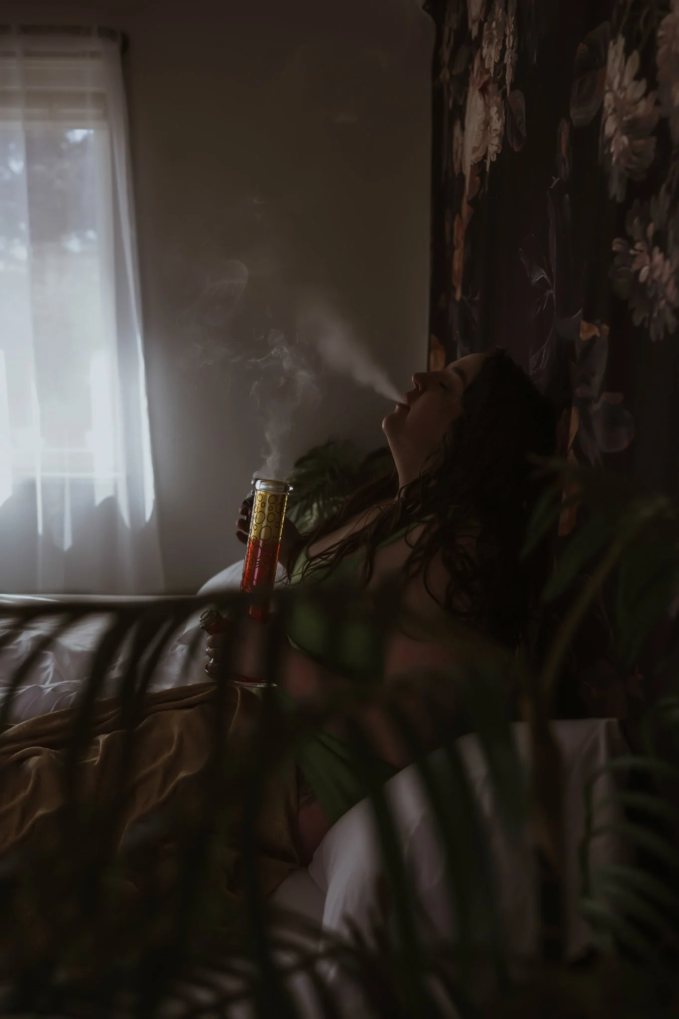 A woman with curly hair reclining on a bed, exhaling smoke from a bong inside a dimly lit room with floral wallpaper and a window with sheer curtains.