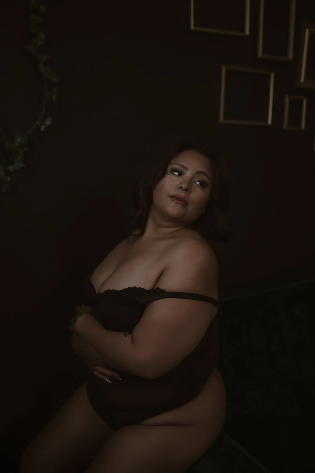 A woman wearing black lingerie sitting with arms crossed in a dimly lit room with black walls and photo frames.