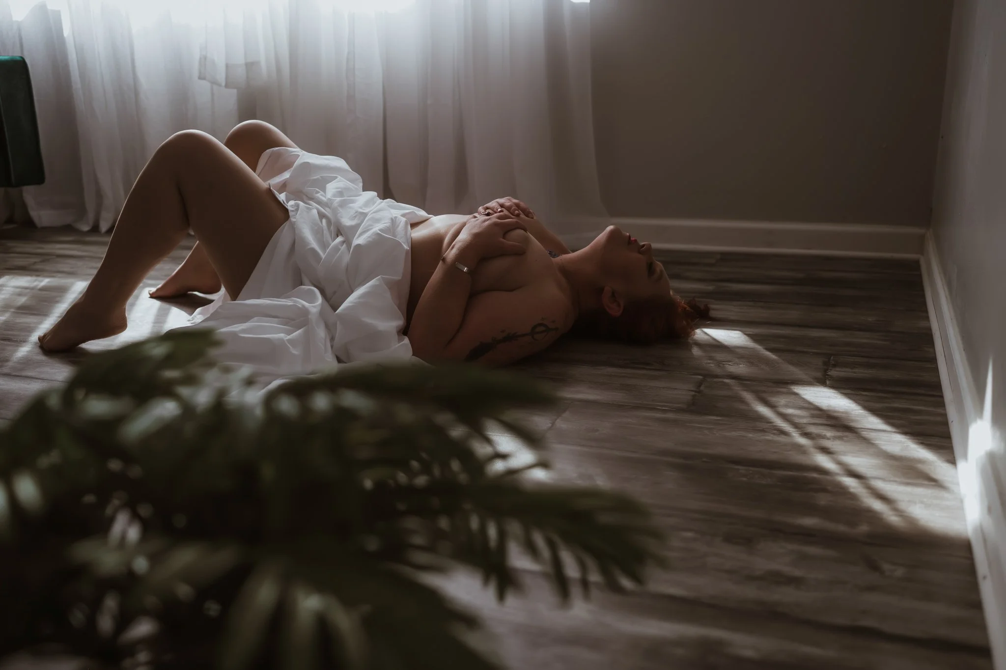 A woman lying on the wooden floor in a dimly lit room, partially covered by a white sheet, with her hand on her chest and her head tilted back.