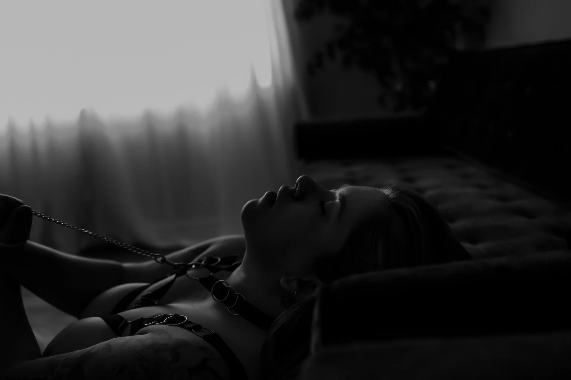 A black and white photograph of a woman lying on her back on a bed, wearing leather straps and a collar with a chain, in a dimly lit room with a window and curtains in the background.