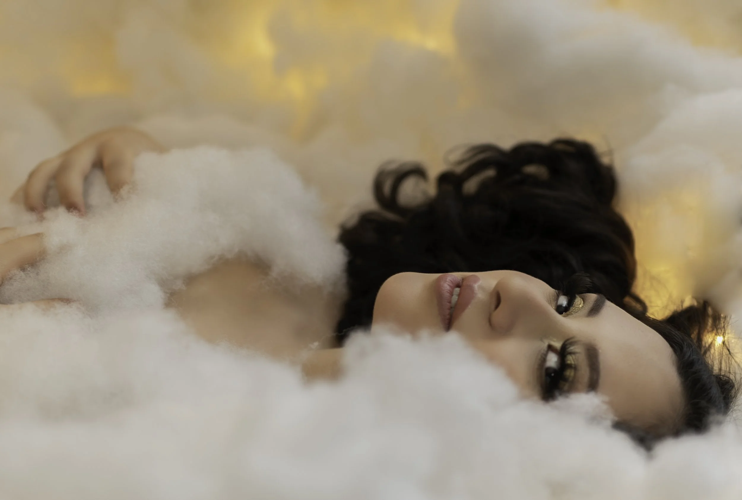 A woman with dark, wavy hair lying among fluffy white clouds or mist, looking at the camera with a gentle expression, and a background of warm, glowing lights.