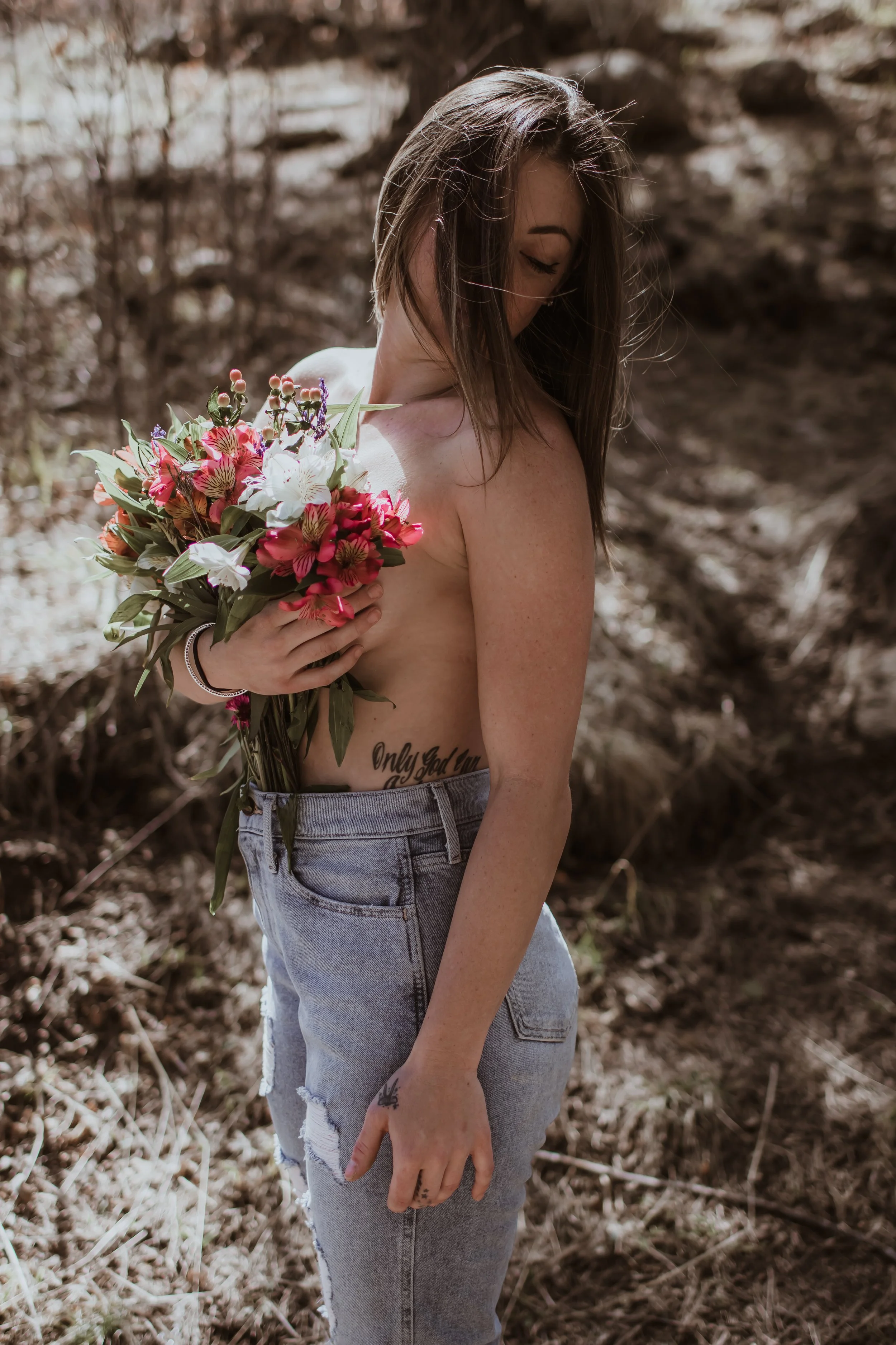 A woman with long brown hair, wearing ripped jeans, topless, holding a bouquet of pink and white flowers, standing outdoors with a blurred natural background.