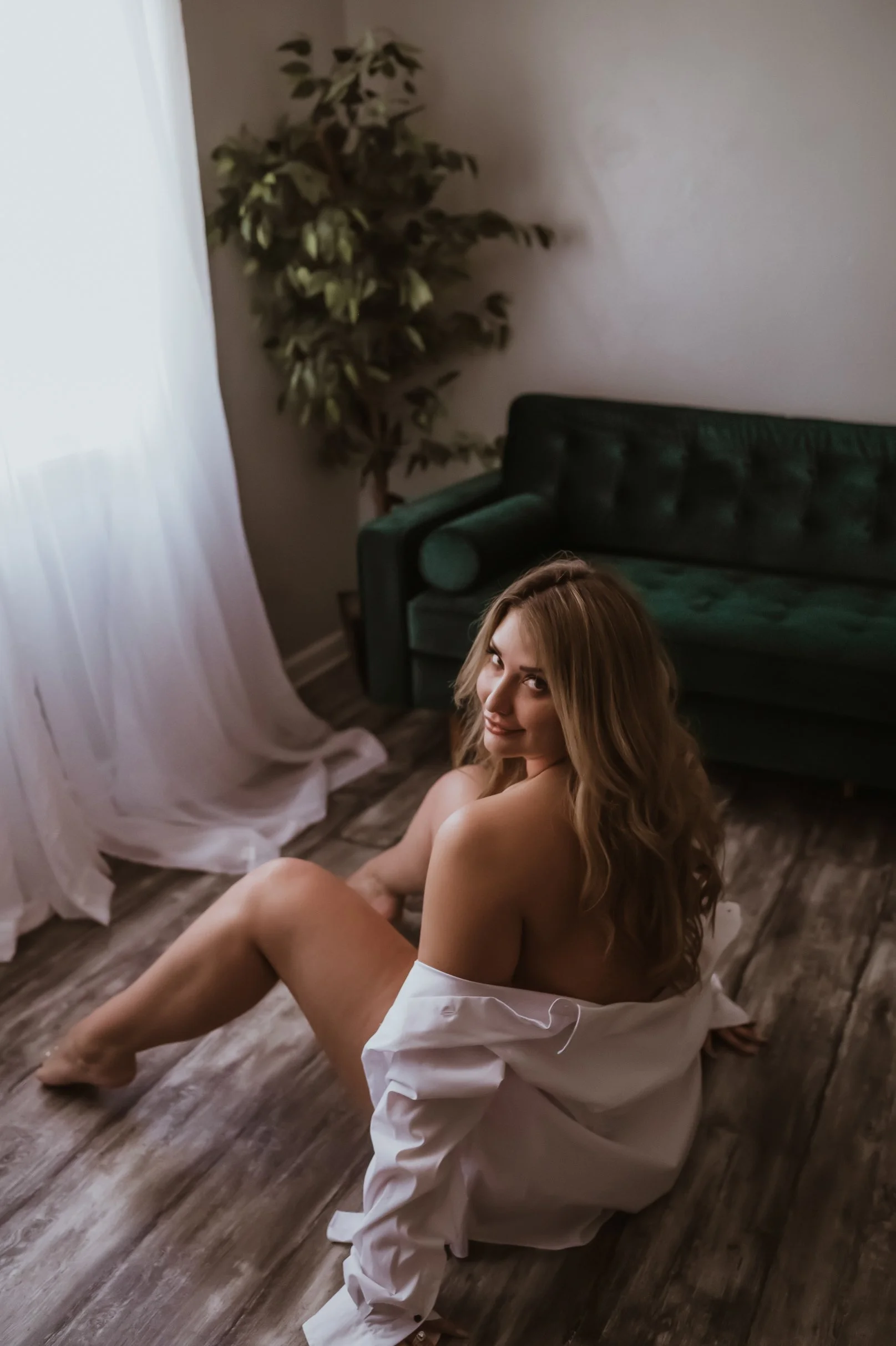 A woman with long wavy hair sitting on a wooden floor near a green couch, smiling at the camera, with a white shirt draped over her shoulders, a plant in the background, and soft natural light coming through white curtains.