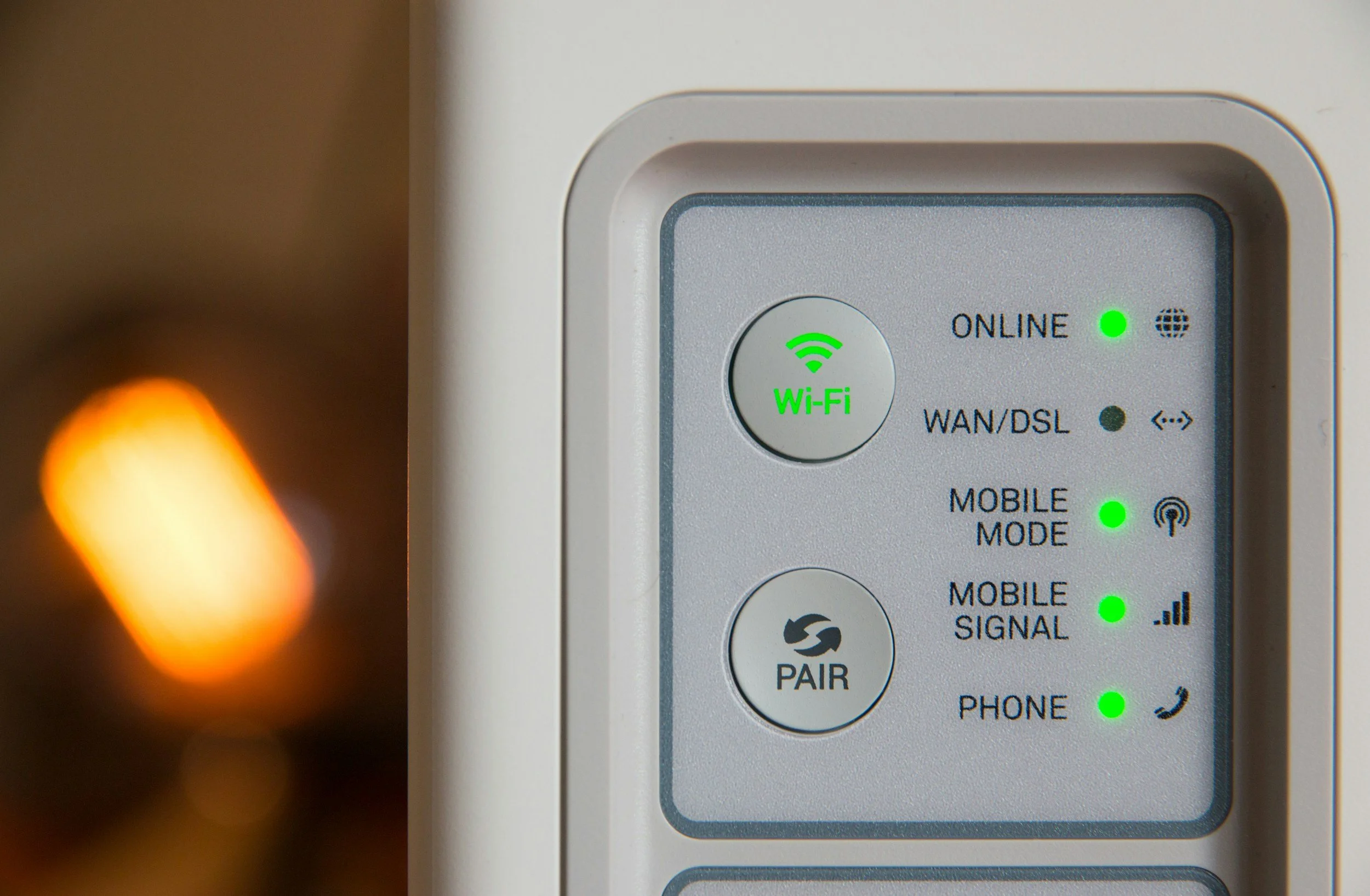 Close-up of a white wireless router with green status lights. The router shows indicators for online status, Wi-Fi, WAN/DSL, mobile mode, mobile signal, and paired device. There is an orange bokeh light out of focus in the background.