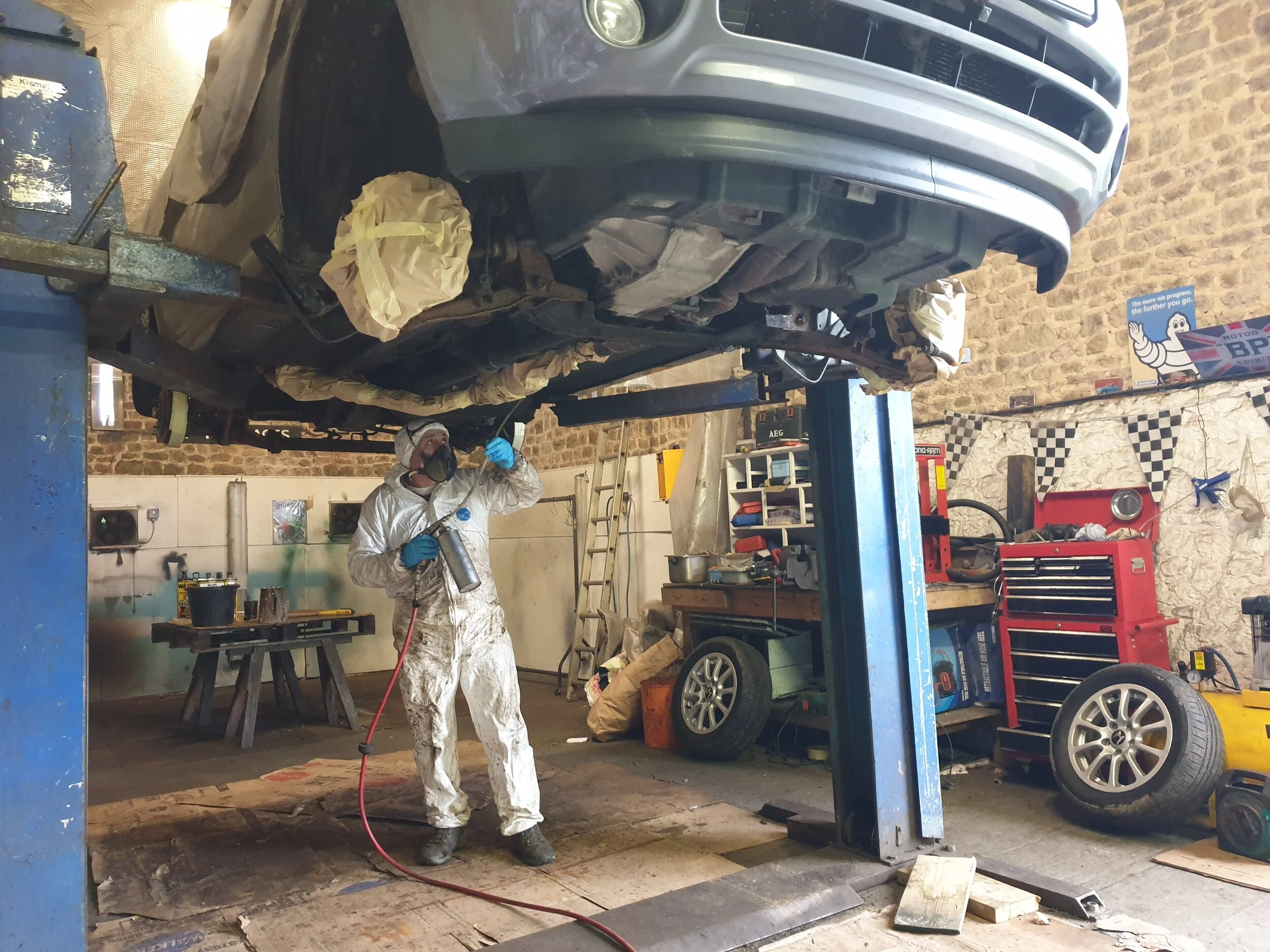 A person in protective gear cleaning or painting a car elevated on a lift in an auto repair shop.