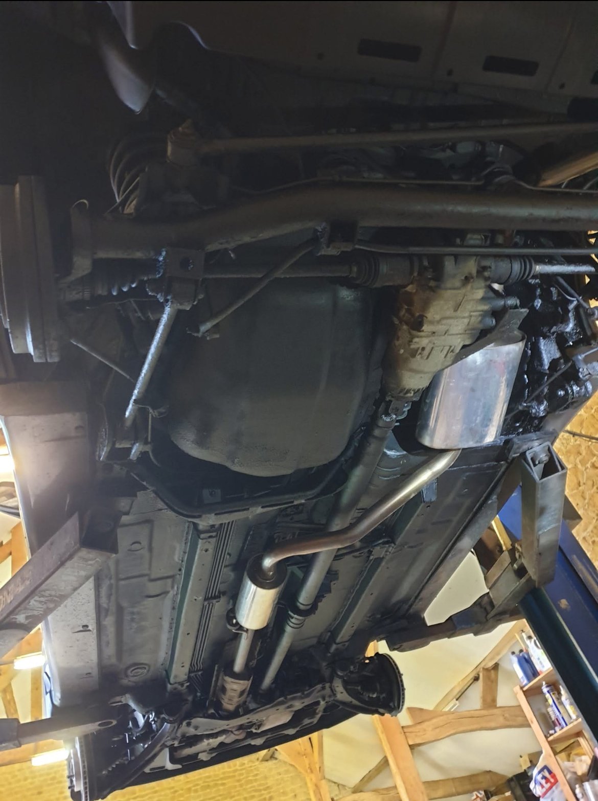 Underside of a vehicle showing the oil pan, transmission, fuel lines, and exhaust system in a workshop.