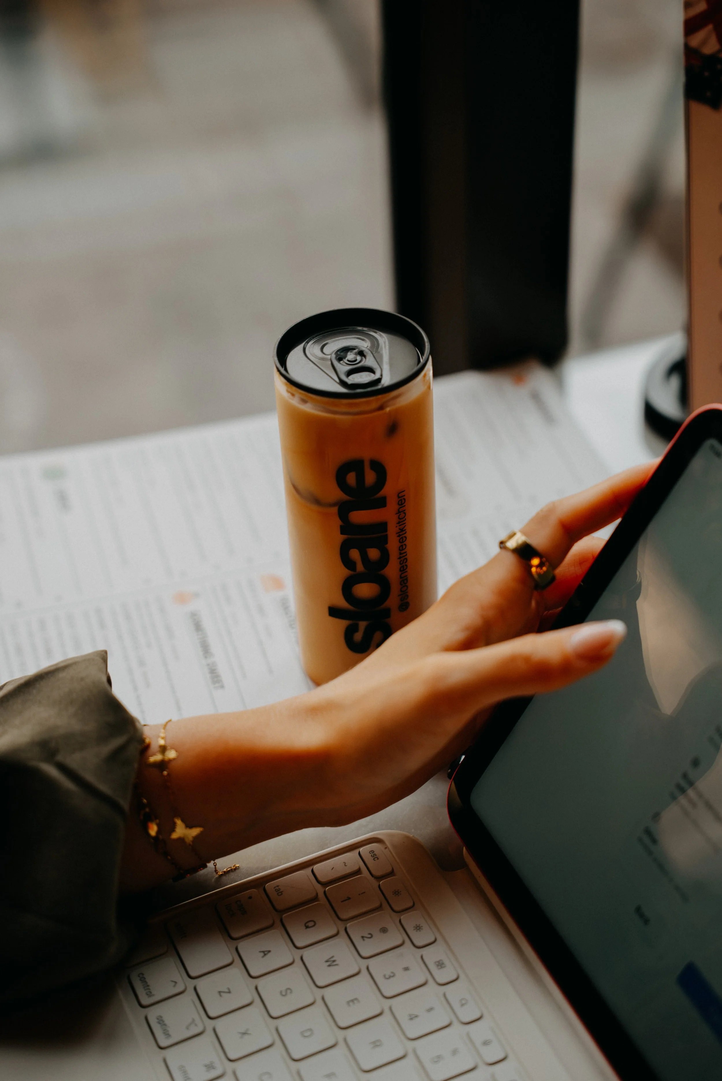 A person with a gold bracelet on their wrist holding a smartphone over a desk with a white keyboard, a newspaper, and a can of an energy drink, with a computer monitor in the background.