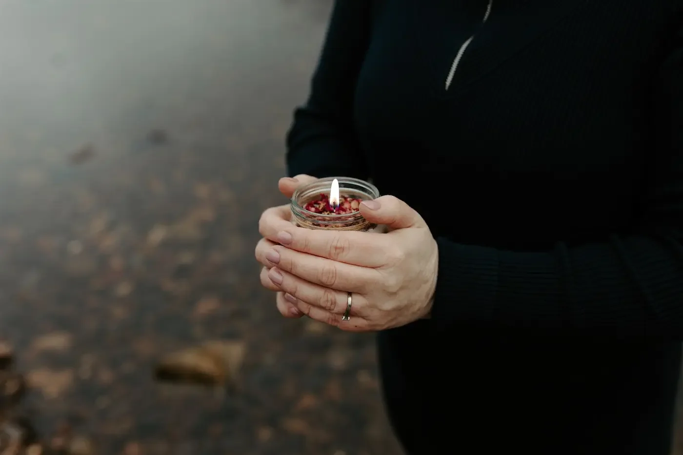 Michelle Lang, Shamanic Practitioner, holding a burning candle outdoors by a shallow river, surrounded by earthy tones during an outdoor ritual.