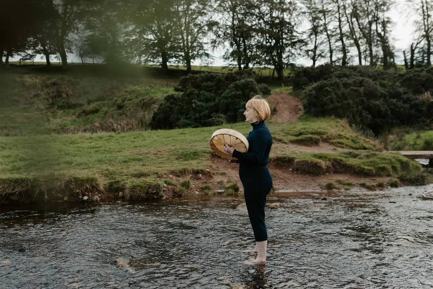 Michelle Lang, Shamanic Practitioner, beating her shamanic drum while standing in a shallow river during a nature immersion near Glasgow.