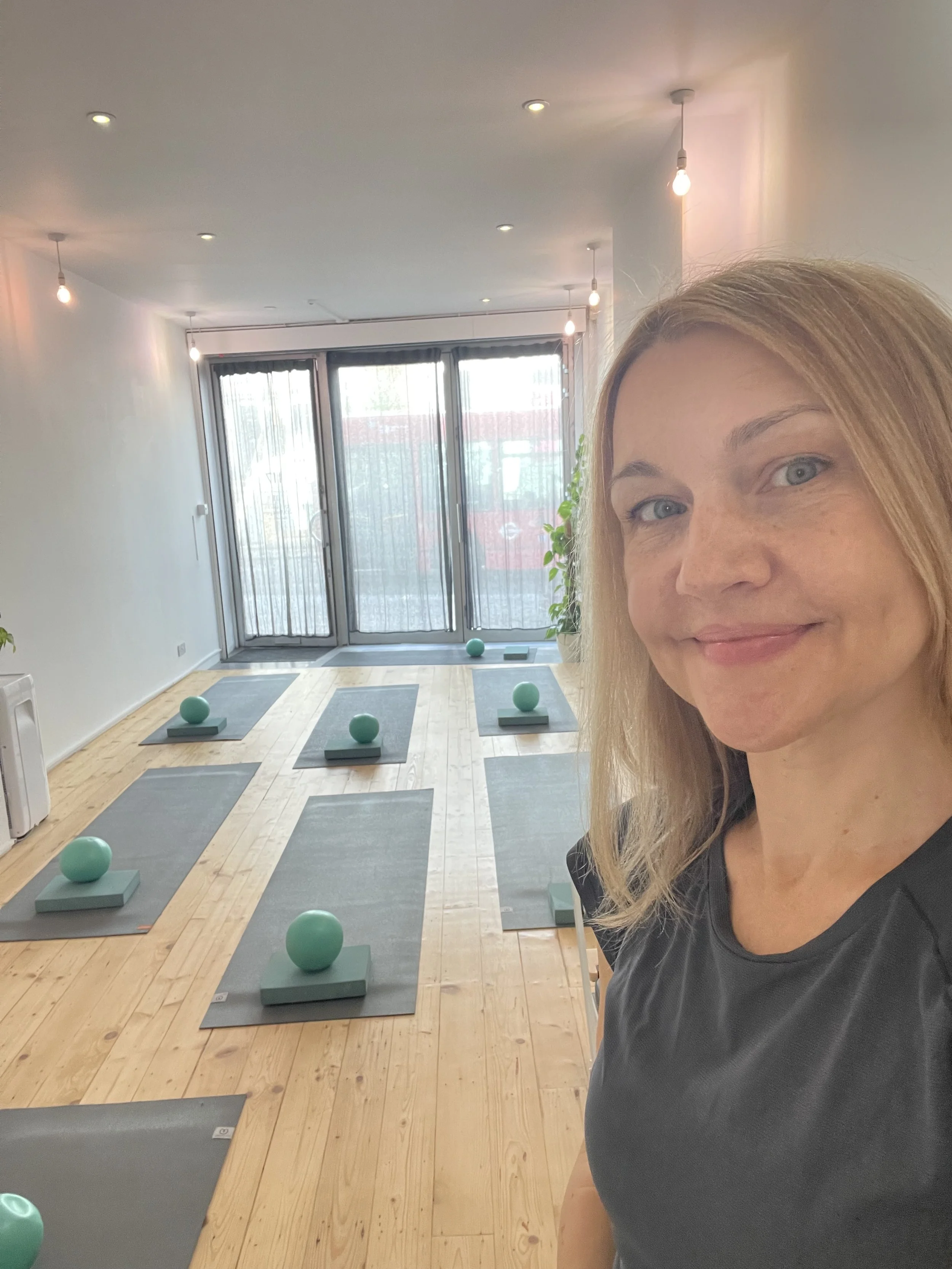 A woman taking a selfie in a yoga or fitness studio with yoga mats and teal-colored exercise balls arranged on the floor, large glass doors with curtains, wooden flooring, and ceiling lights.