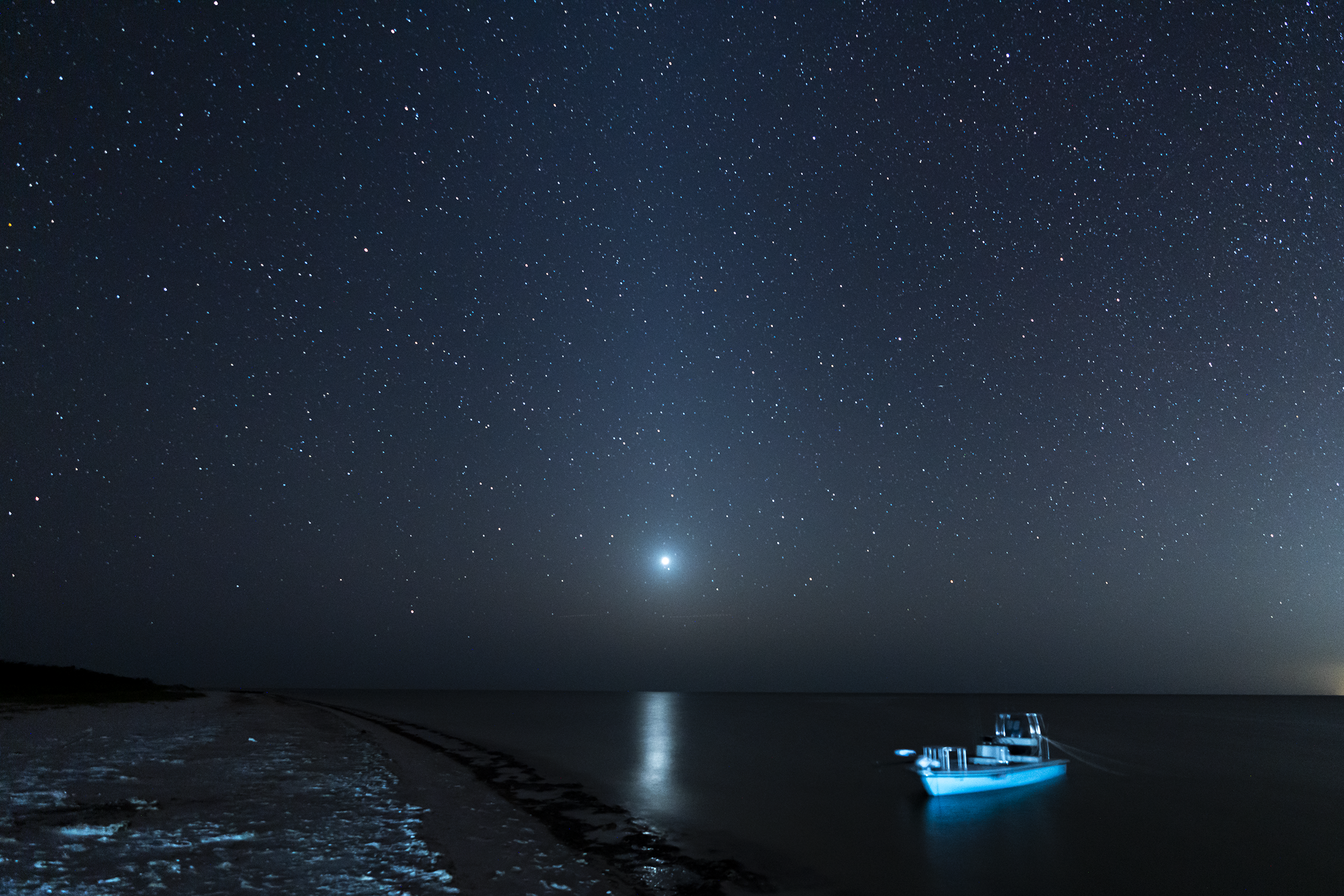 Night sky filled with stars over a shoreline with a boat illuminated in blue light