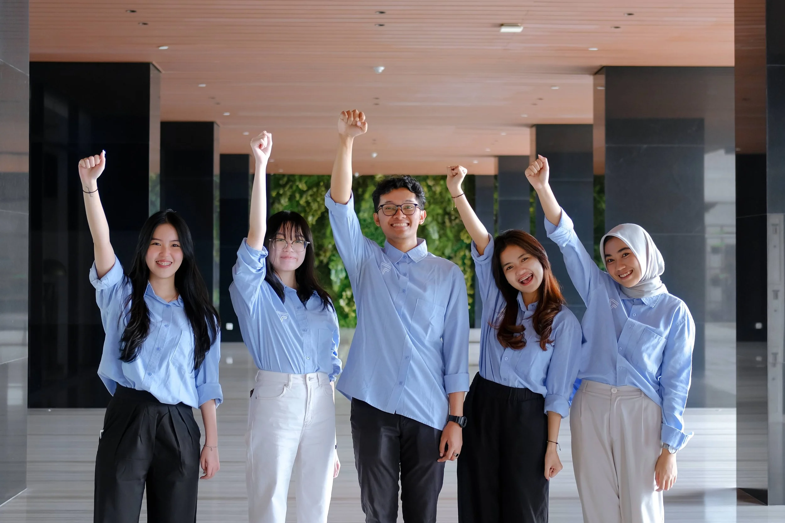 Group of five young adults standing indoors, celebrating with raised fists, wearing light blue shirts, smiling, and looking happy.