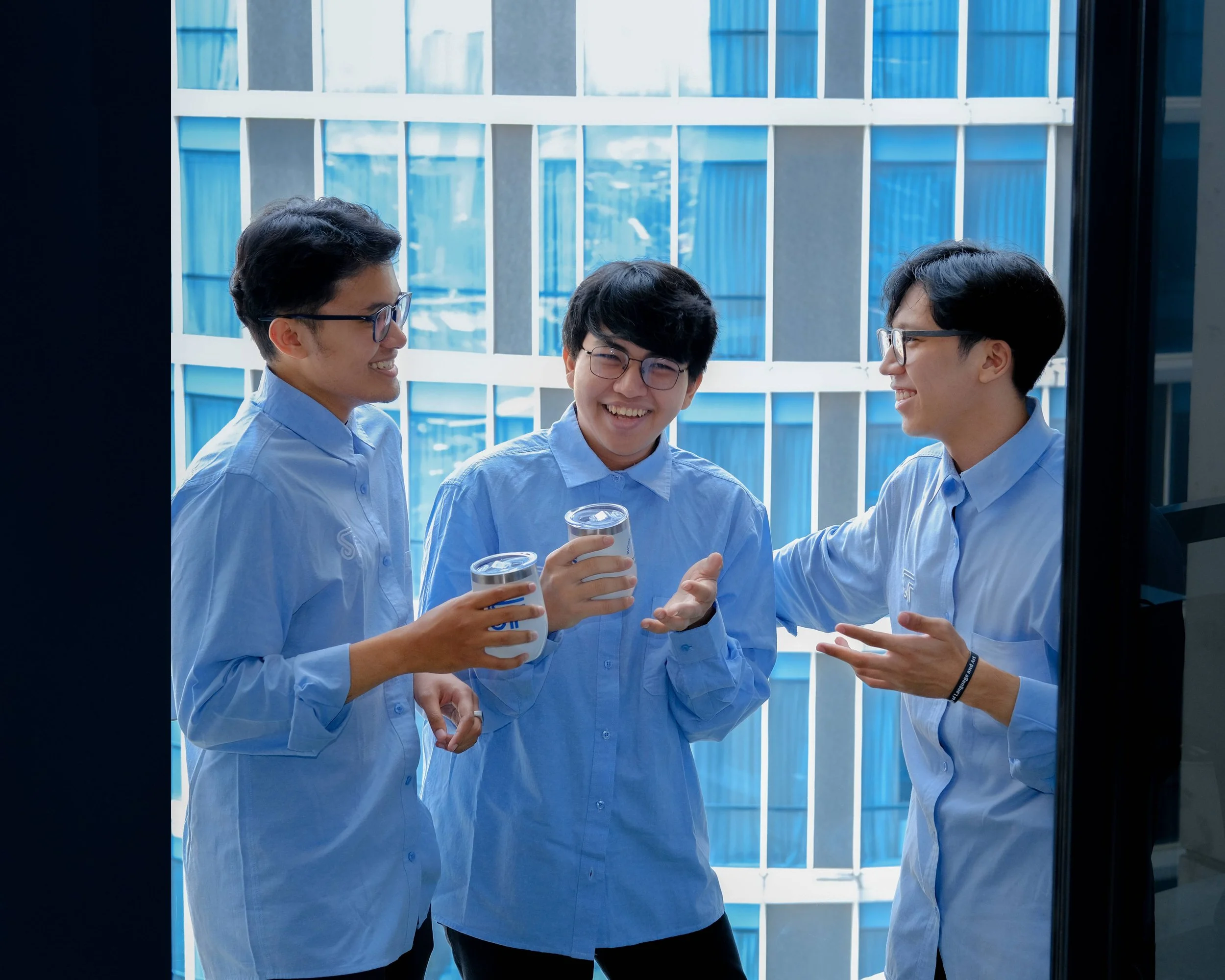 Three young men with glasses wearing light blue shirts talking and smiling, holding drinks, standing outside near a modern glass building.