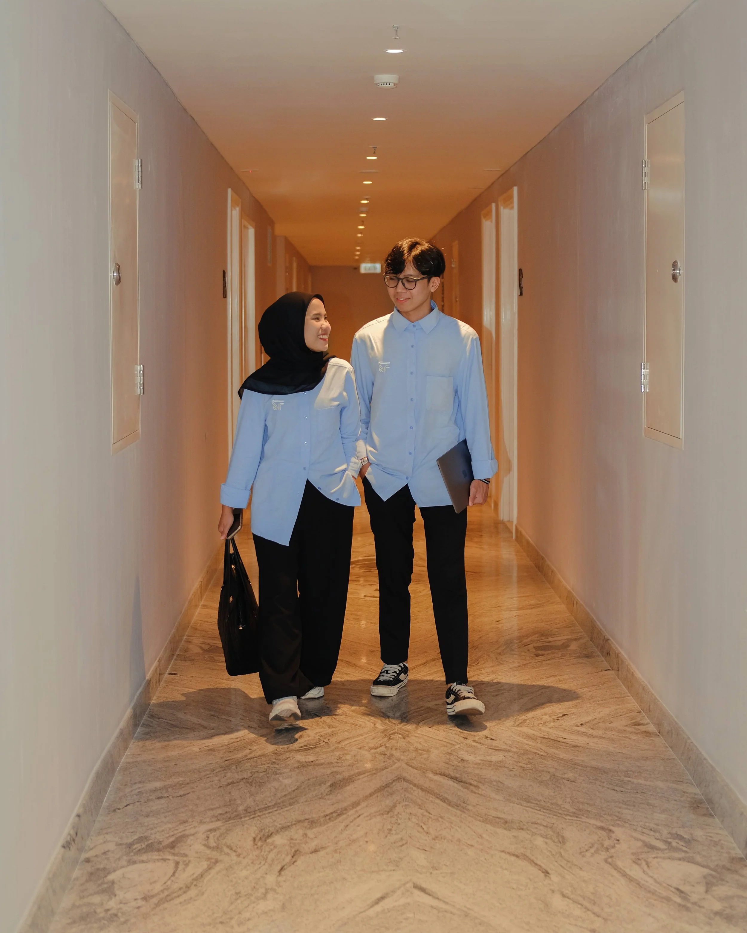 Two colleagues, a woman wearing a hijab and a man, walking and talking down a corridor in an office building, both dressed in light blue shirts and dark pants.