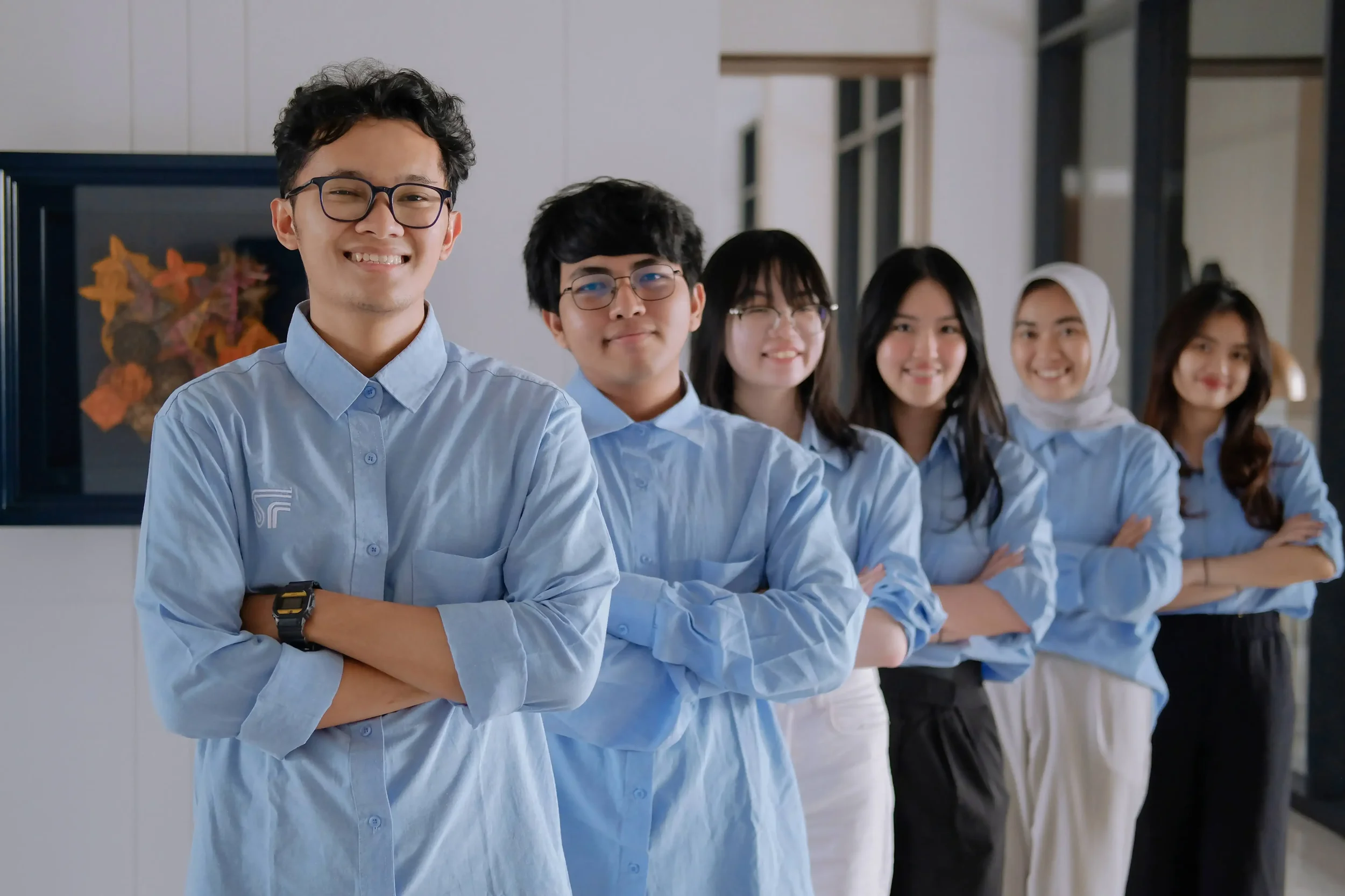 A group of six diverse young adults standing in a line indoors, smiling, wearing light blue shirts, with arms crossed or hands at their sides. The setting appears to be a professional or academic environment.