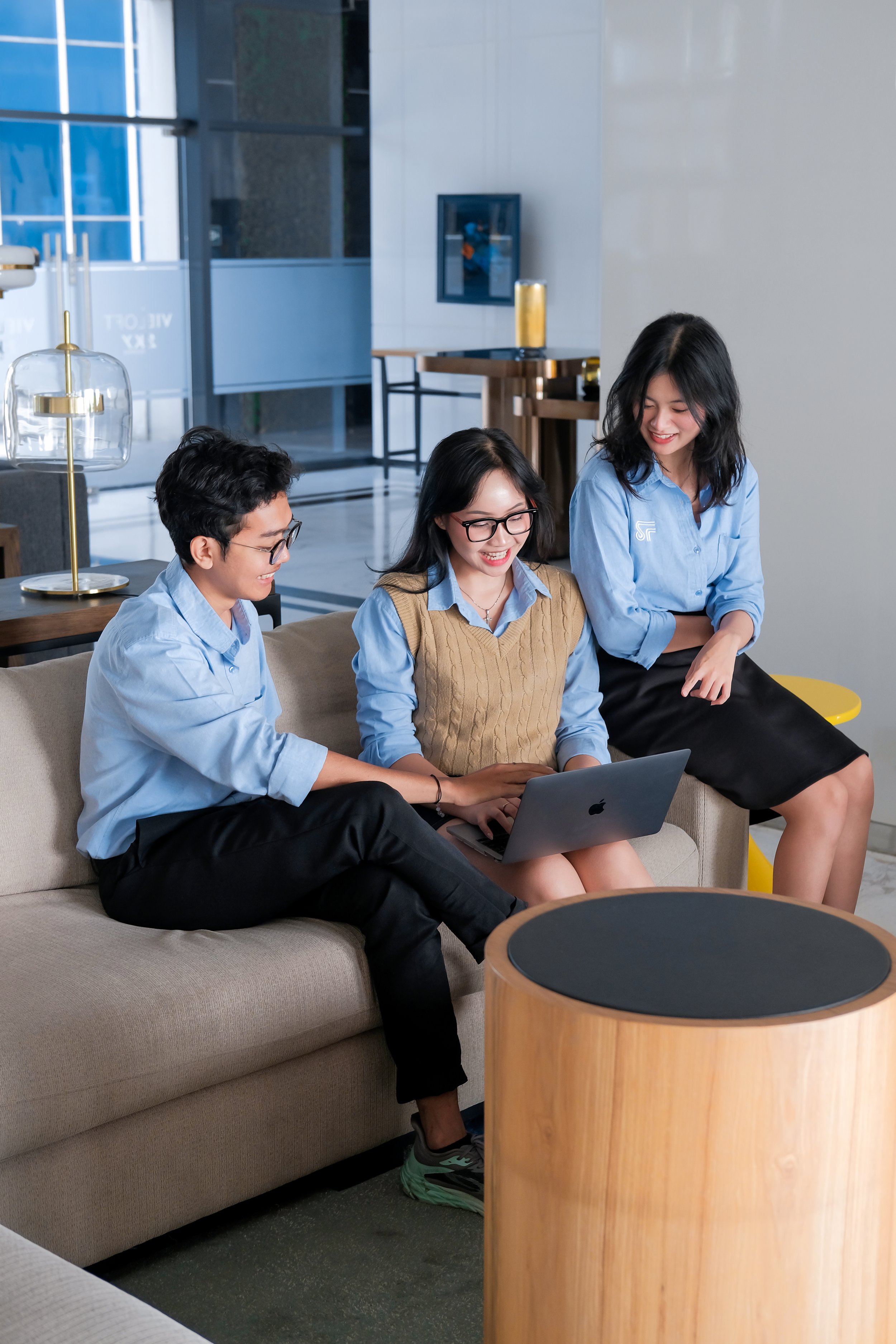 Three colleagues sitting on a beige sofa in a modern office lounge, smiling and looking at a laptop, with a small table in front of them.