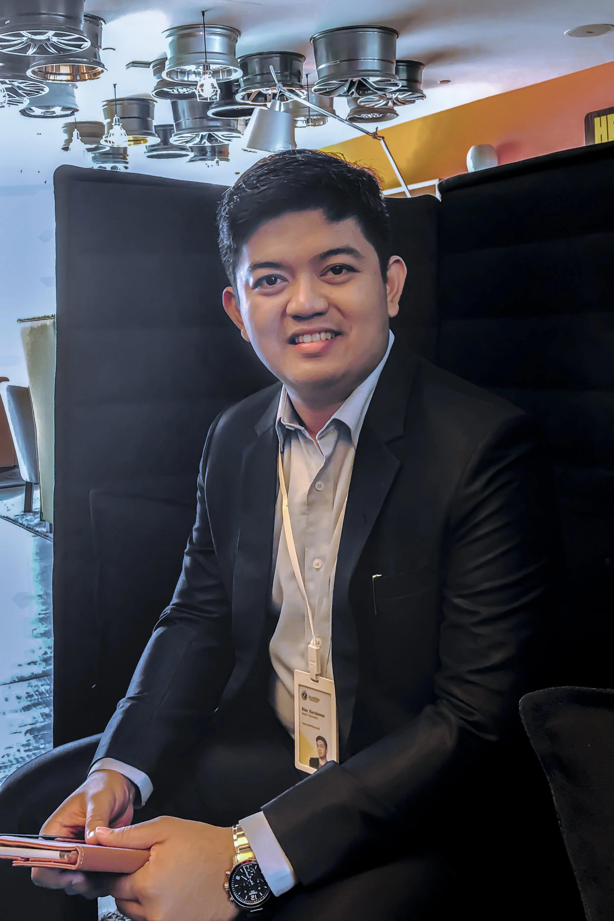 Person sitting with a smile, wearing a suit and lanyard, holding a smartphone, in a modern indoor setting.