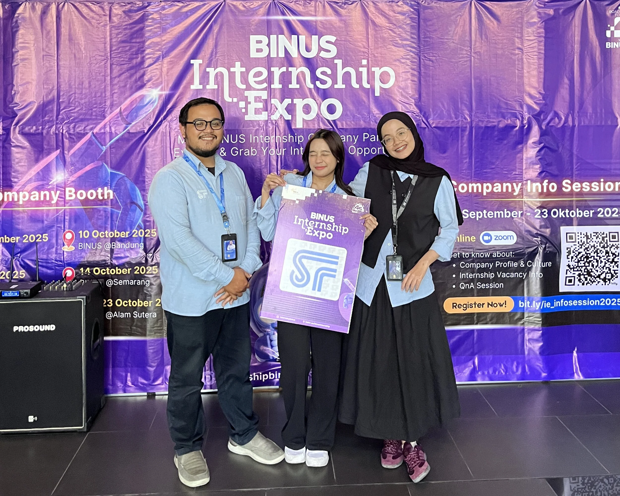 Three people standing in front of a purple banner at the BINUS Internship Expo. The person in the middle is holding a large purple sign. All three are smiling and wearing conference badges.