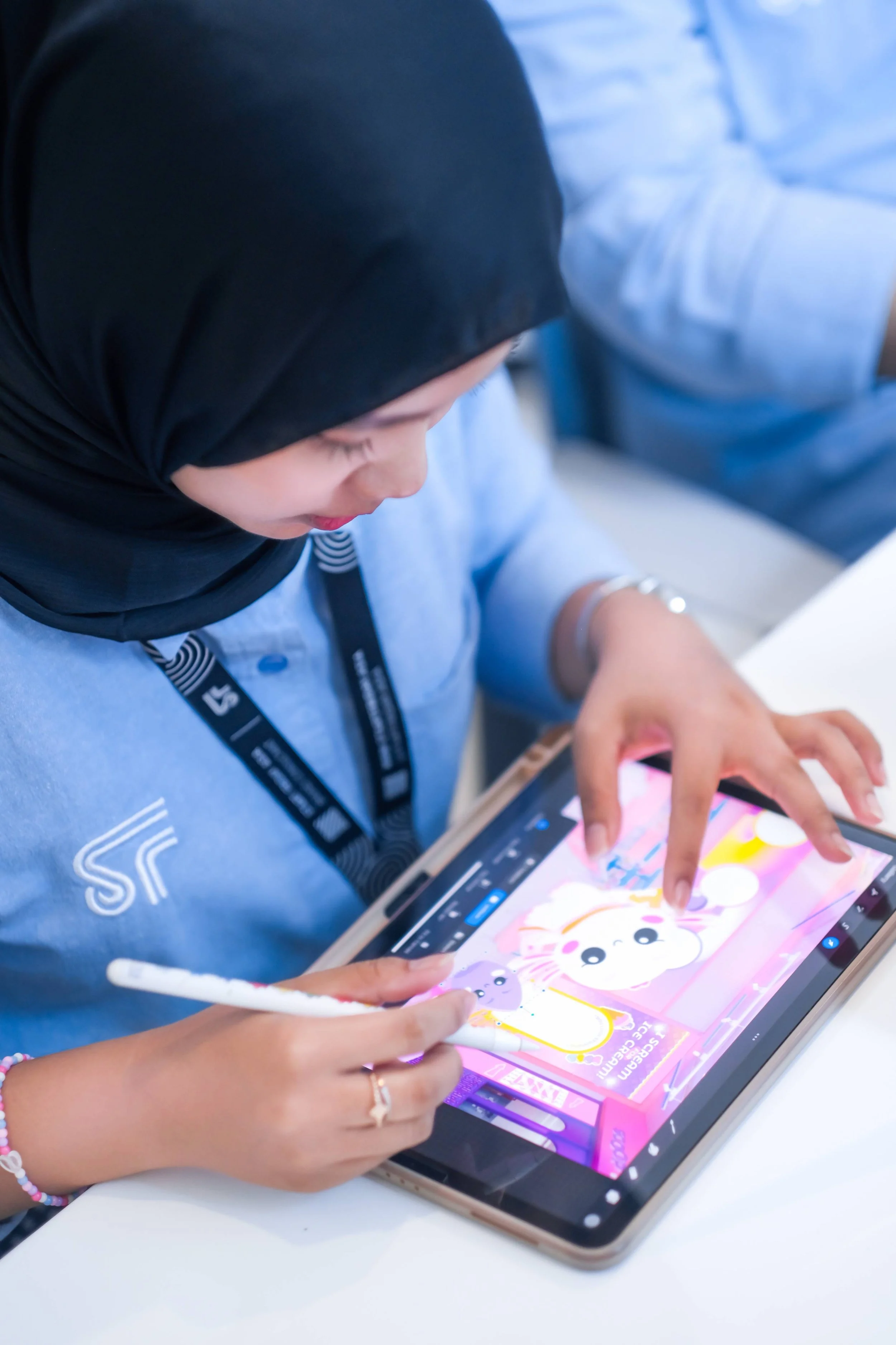 A woman wearing a black hijab and a blue uniform is drawing or editing a colorful cartoon on a tablet with a stylus pen. She is seated at a white table.