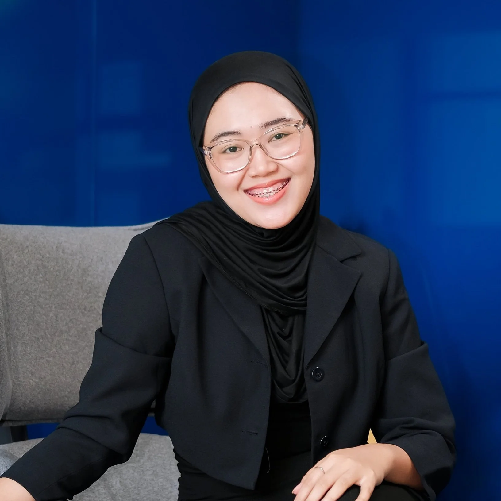 Person wearing a black hijab and suit, smiling, sitting on a gray chair against a blue background.