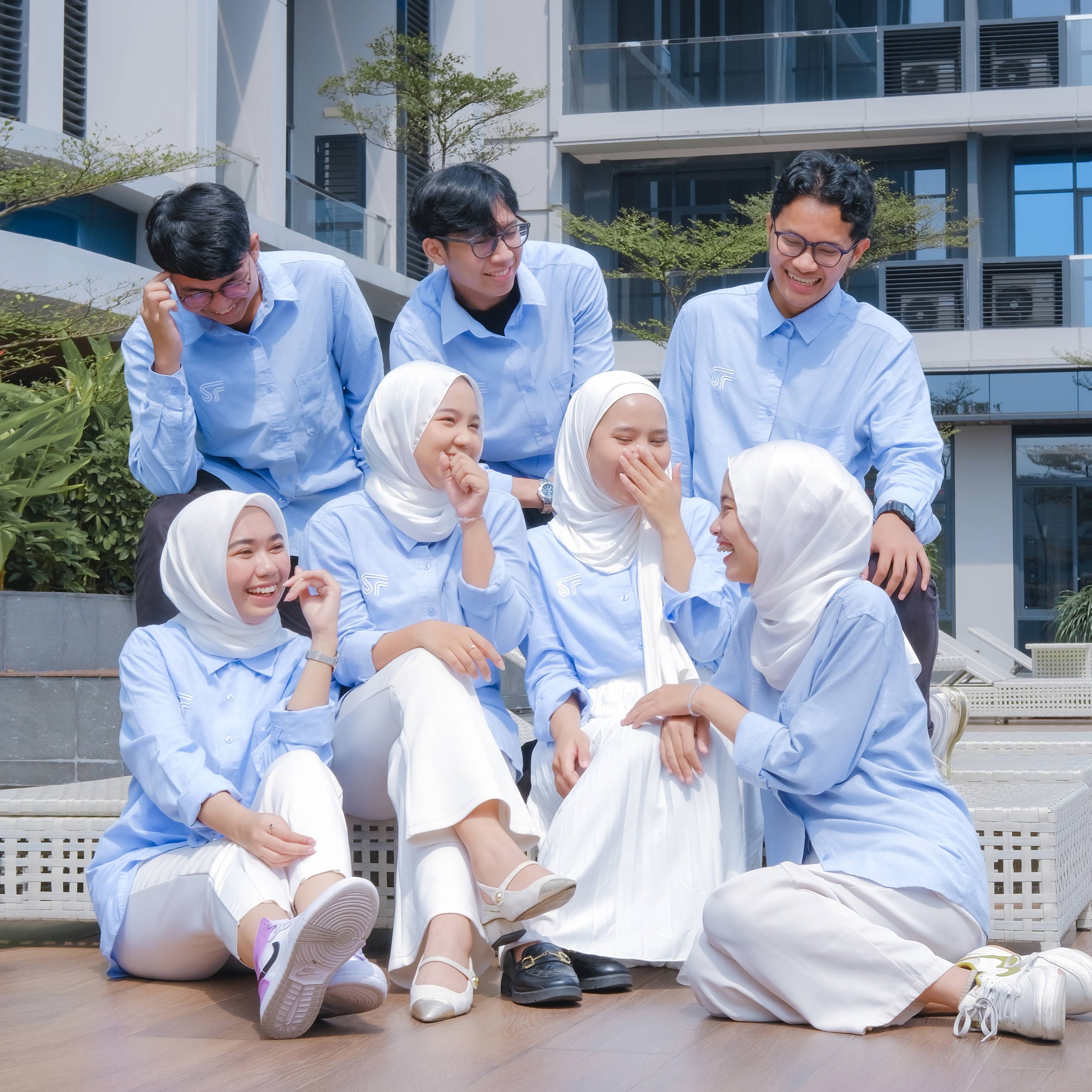 Group of young people wearing matching light blue shirts, laughing and sharing a joyful moment outdoors in front of a modern building with greenery.