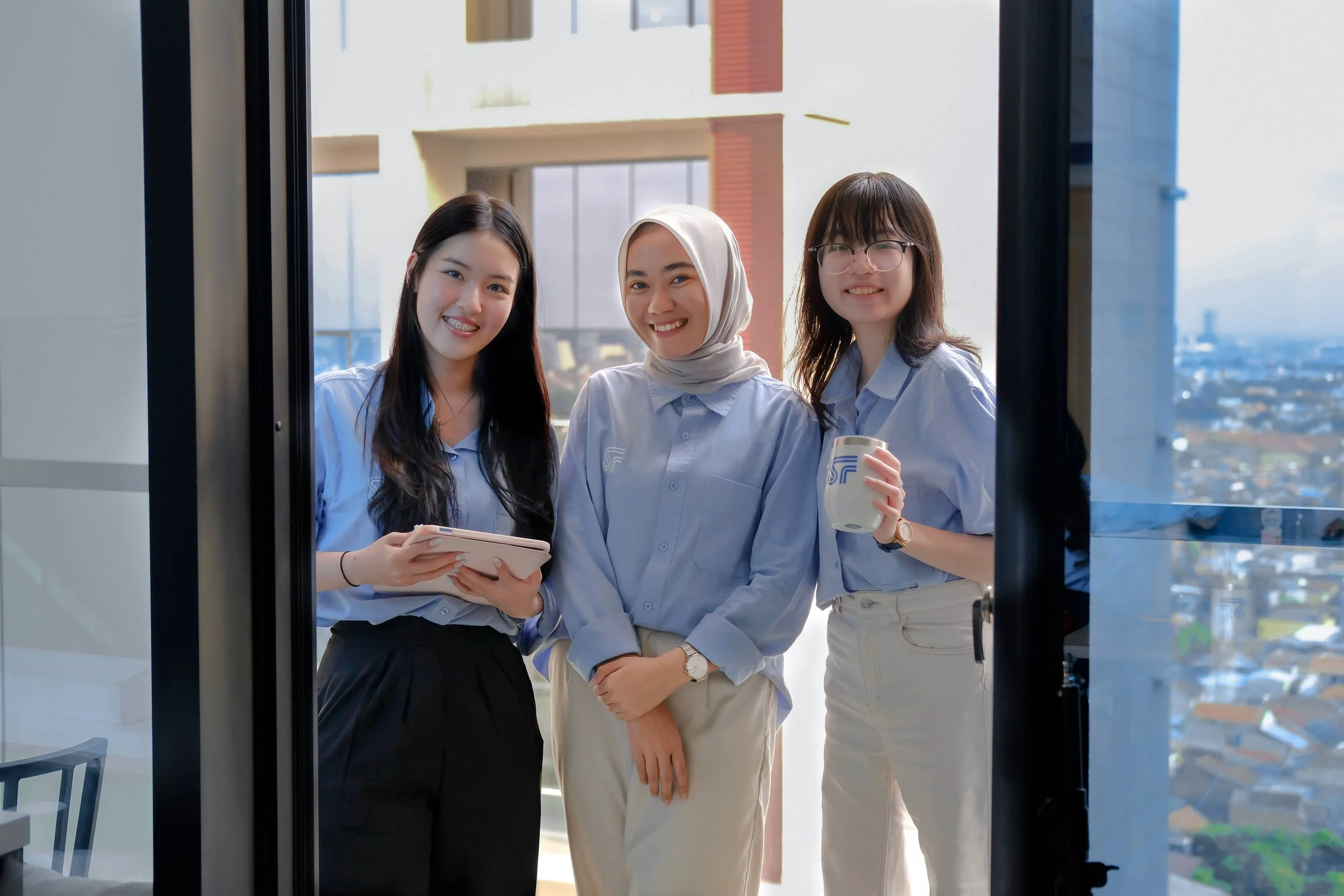 Three women in light blue shirts standing outside an office window, smiling and holding a folder, a mug, and a tablet, with a cityscape background.