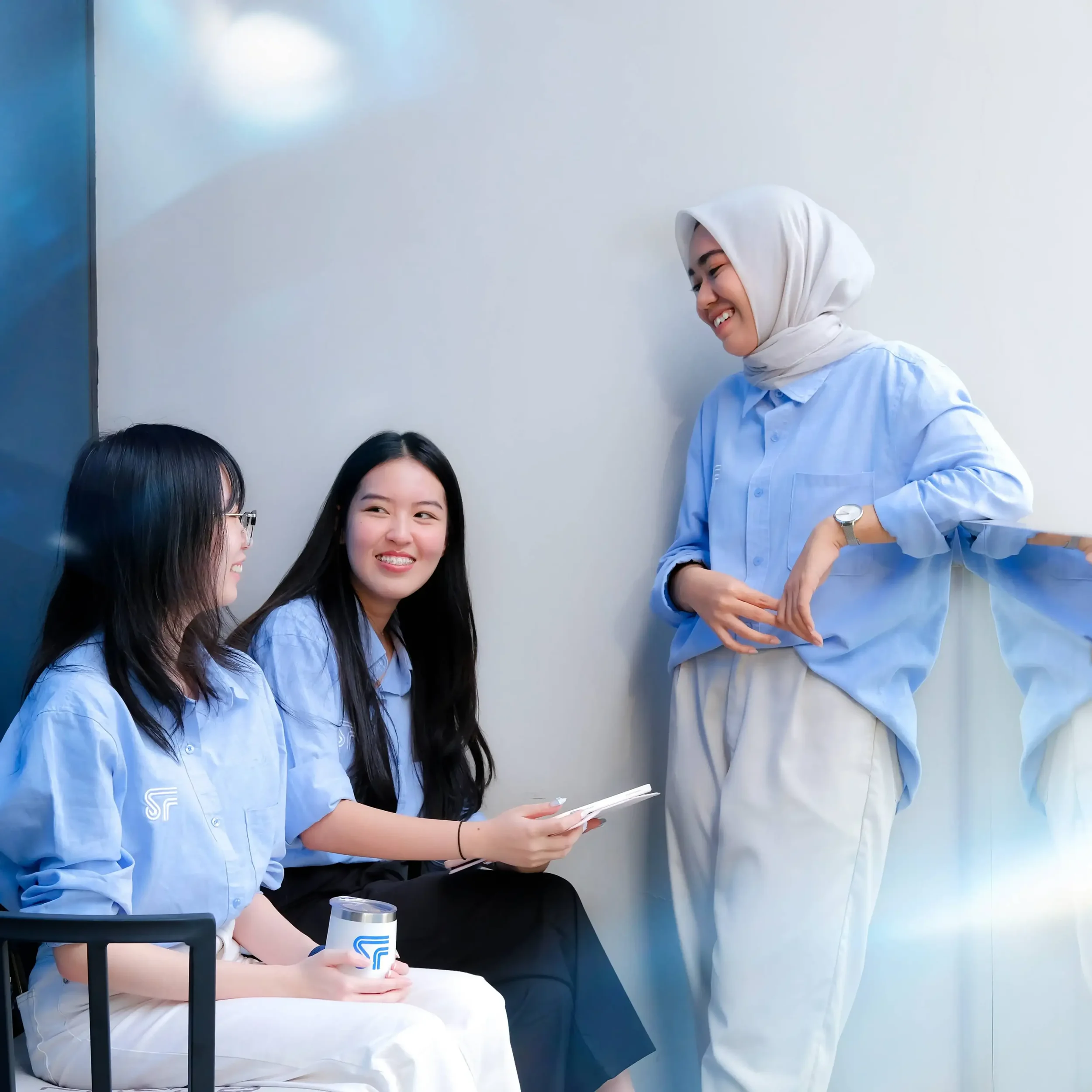 A smiling woman with long dark hair, wearing glasses and a light blue shirt, sitting at a table with a white disposable cup in her hand. She is looking at a standing woman in a hijab and blue shirt, who is holding a paper and engaging in conversation