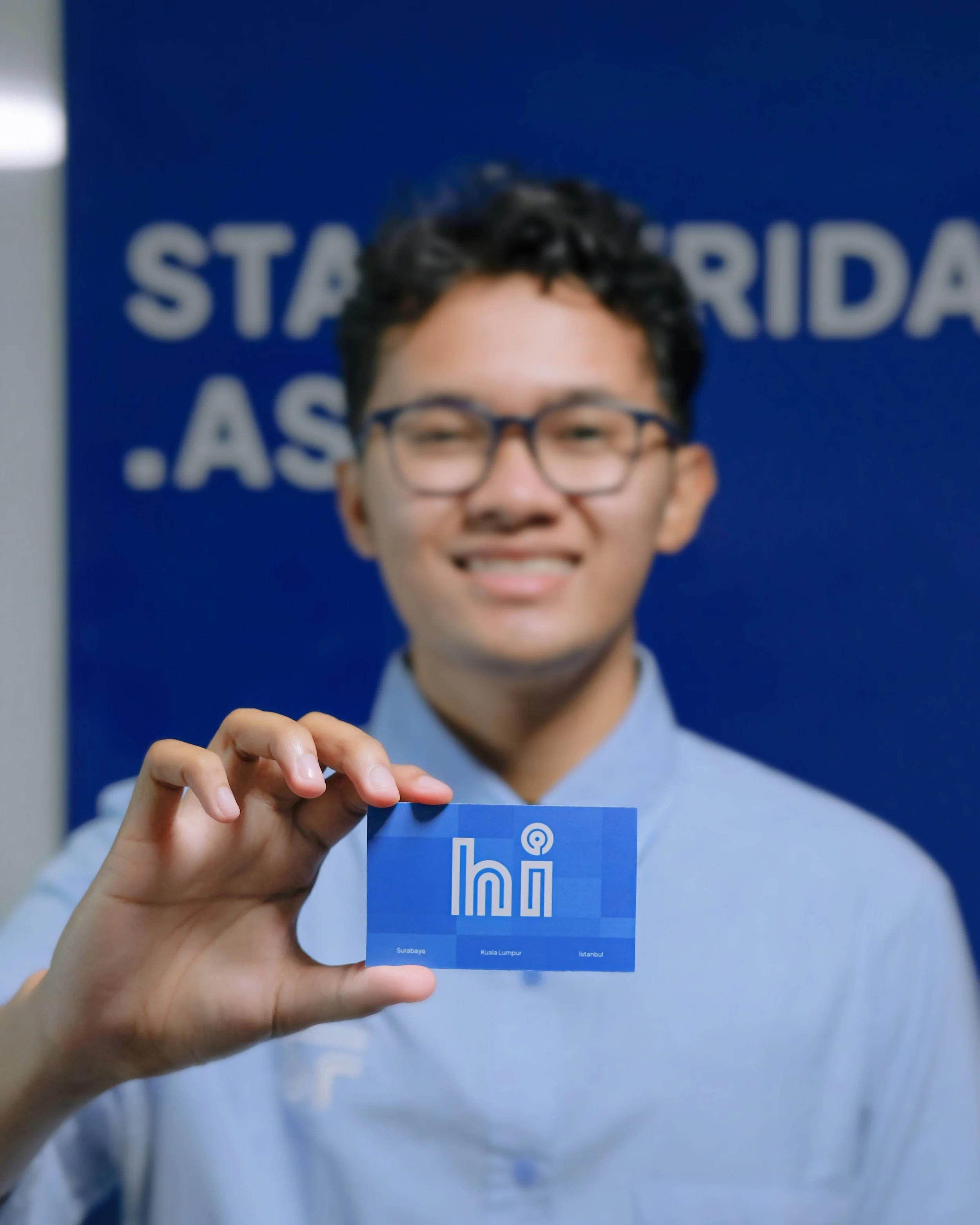 A young man with glasses smiling and holding a blue card saying 'hihi' with the cities Surabaya, Kuala Lumpur, and Istanbul written on it, standing in front of a blue background with similar text.