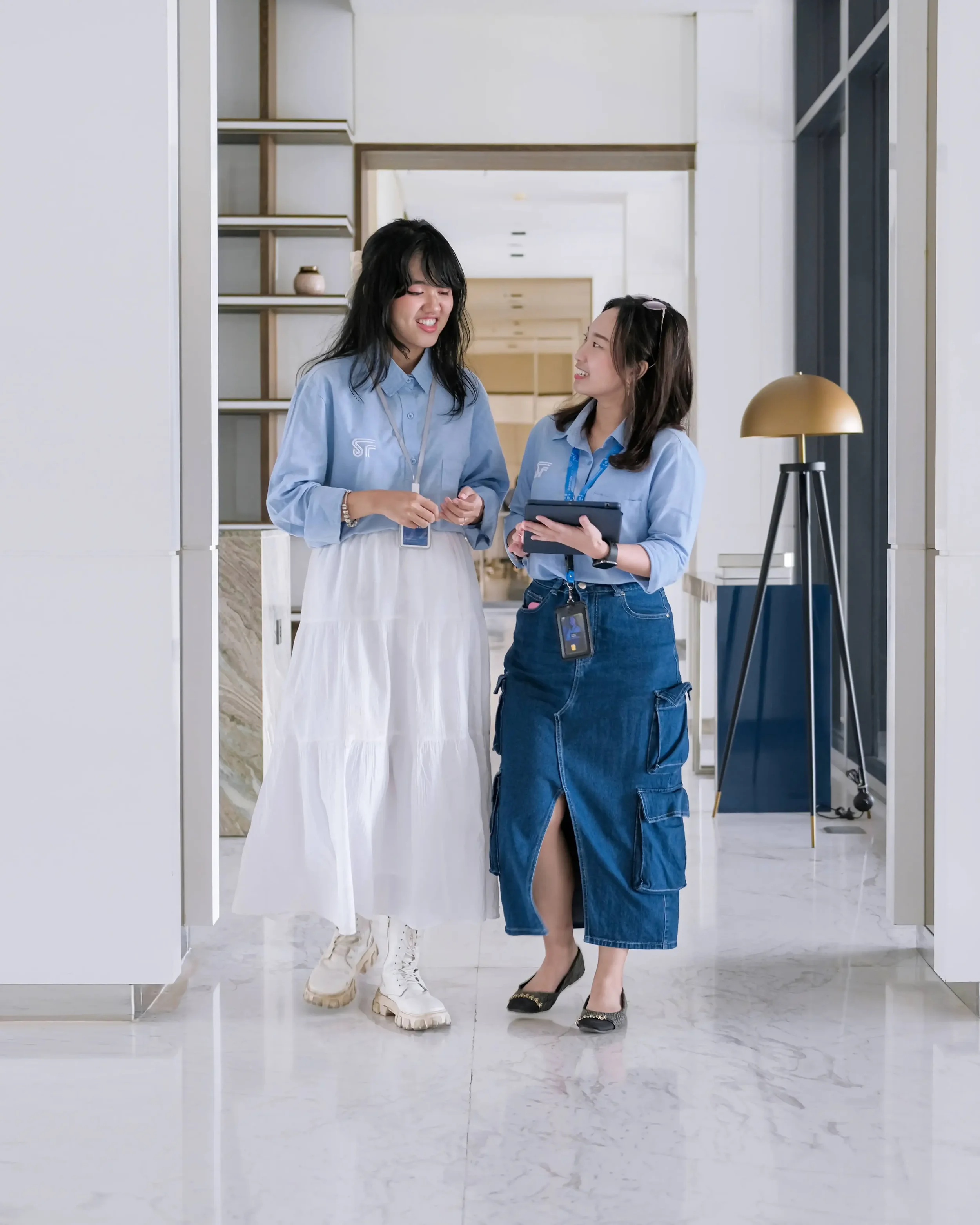 Two women in a bright modern space are smiling at each other. One woman wears a light blue blouse and a long white skirt, and the other wears a light blue shirt and dark blue jeans with a high slit, holding a tablet. They are in a stylish interior wi