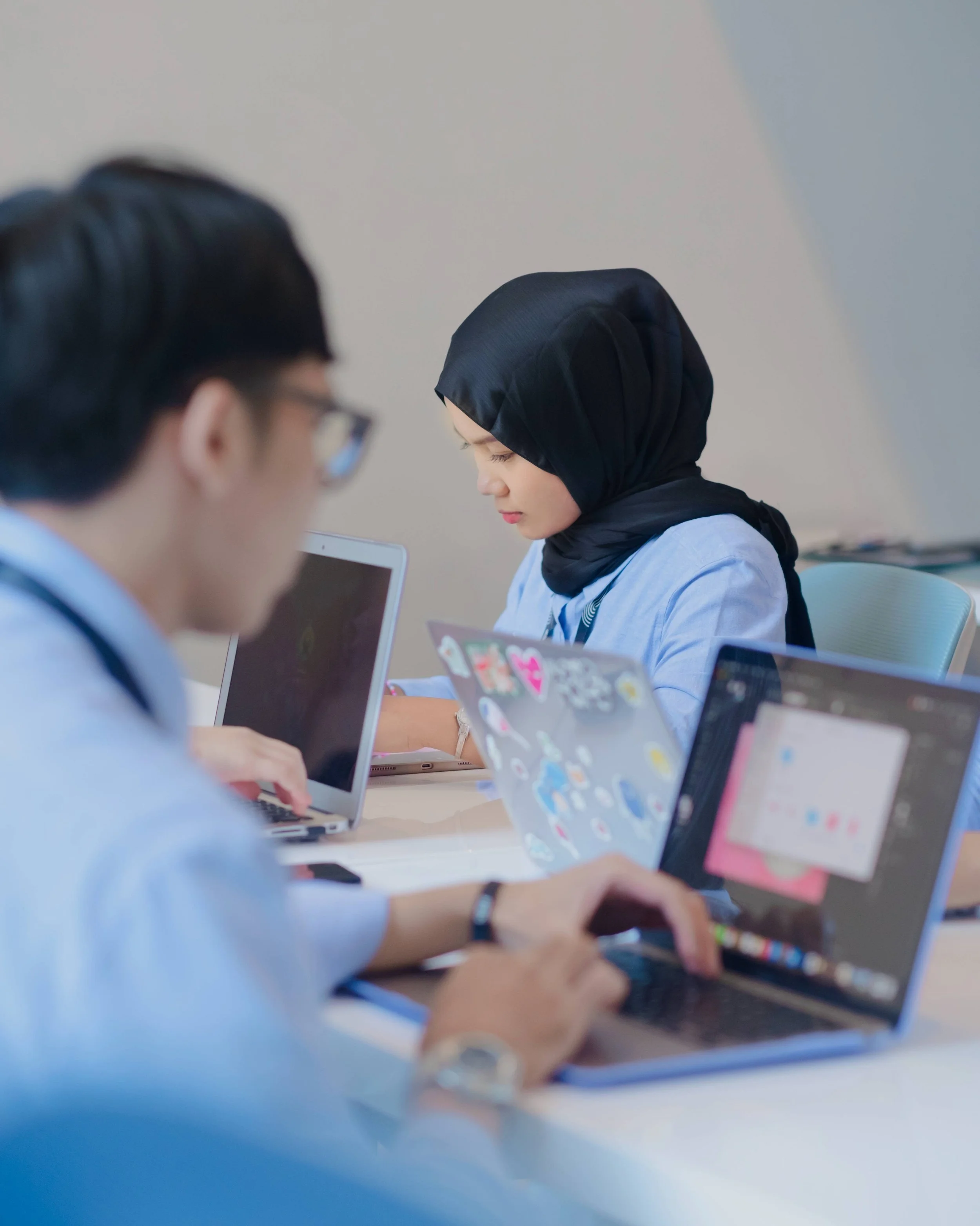 Two people working on laptops at a desk, one wearing glasses and the other wearing a black hijab, in a professional setting.
