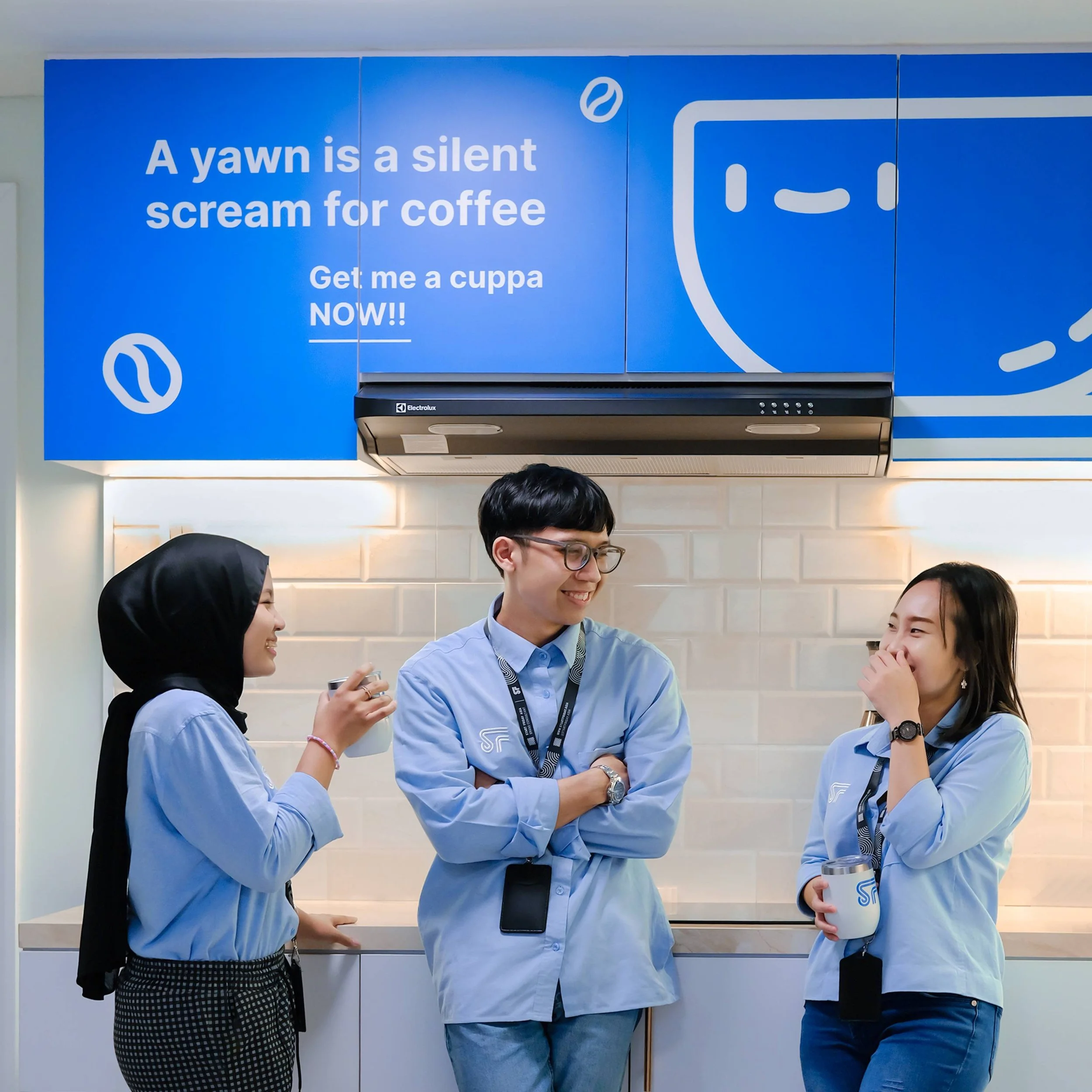 Three colleagues in light blue shirts standing and laughing in a coffee break area, with a blue sign above that reads 'A yawn is a silent scream for coffee' and shows smiling cookie and coffee icons.