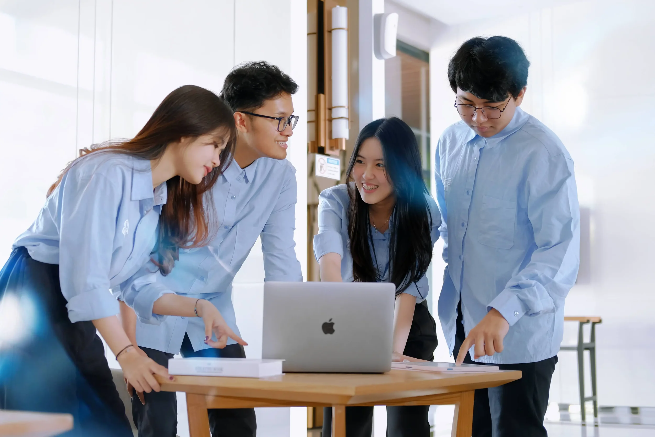 Five young people gathered around a table, looking at a laptop screen, collaborating in a modern, bright office setting.