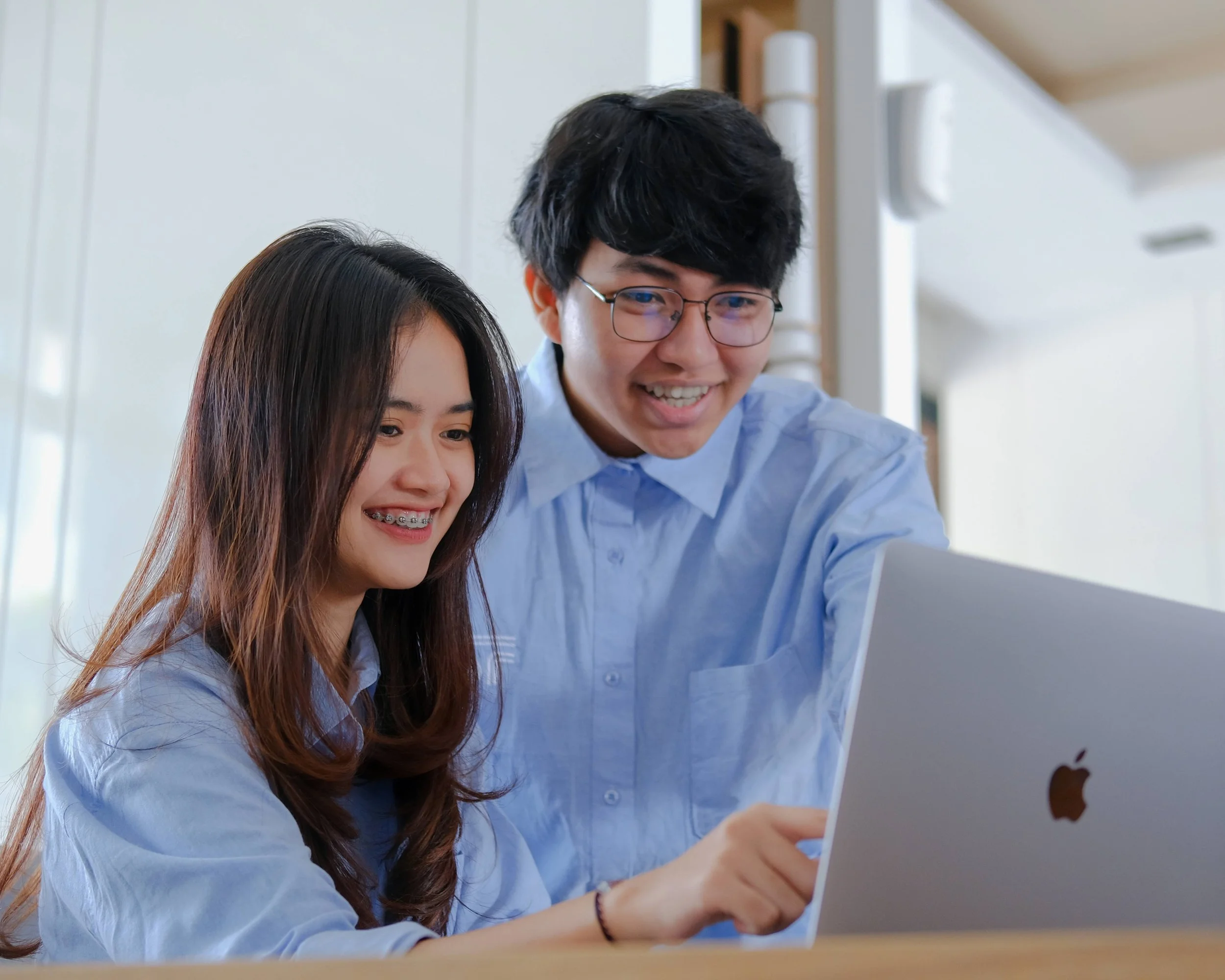 Two young people, a woman and a man, smiling and looking at a laptop computer in a bright indoor setting.