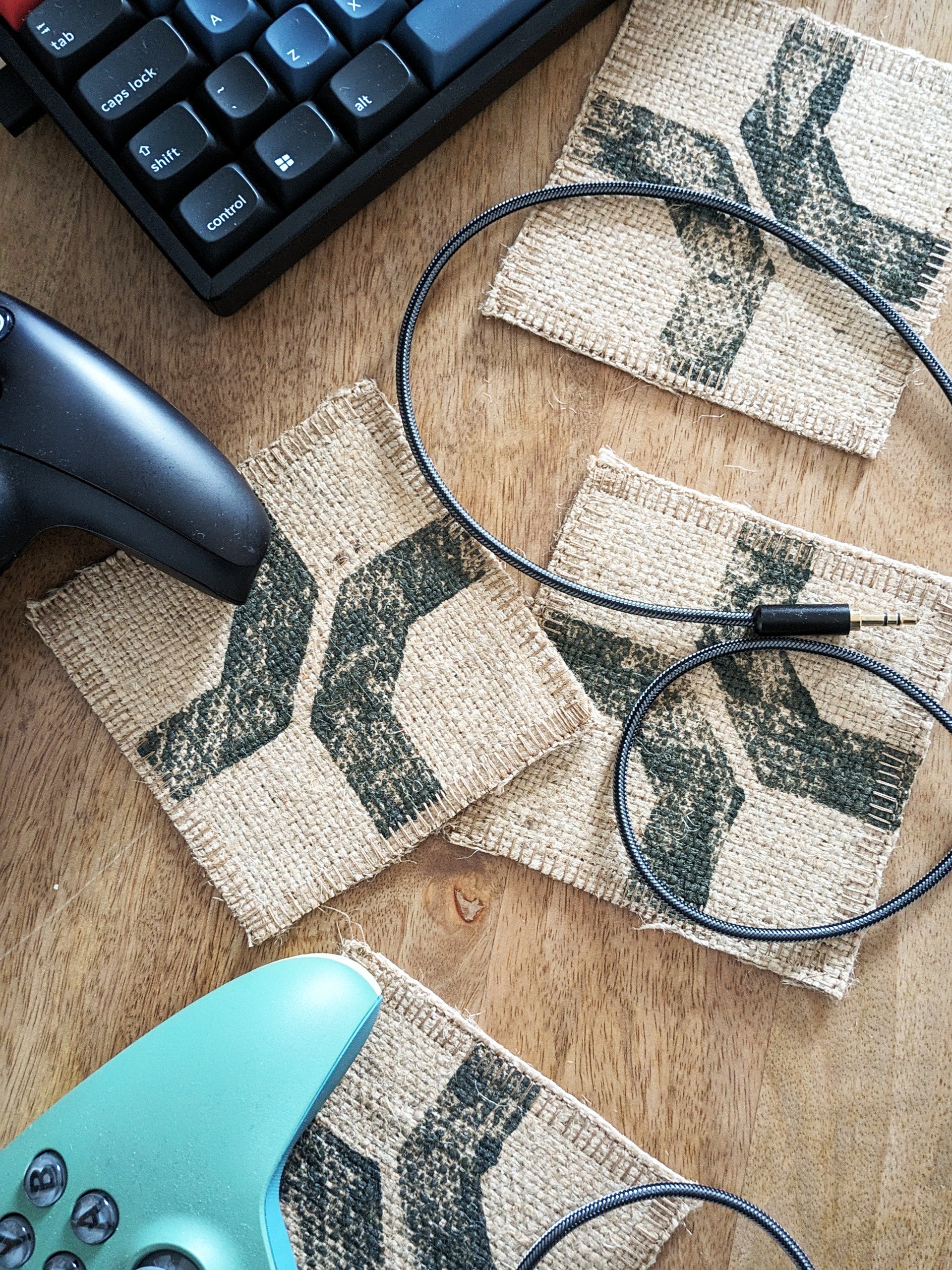 set of 4 coasters on desk with grunge-effect, cross shaped dark khaki print on raw jute, with videogame controllers and keyboard