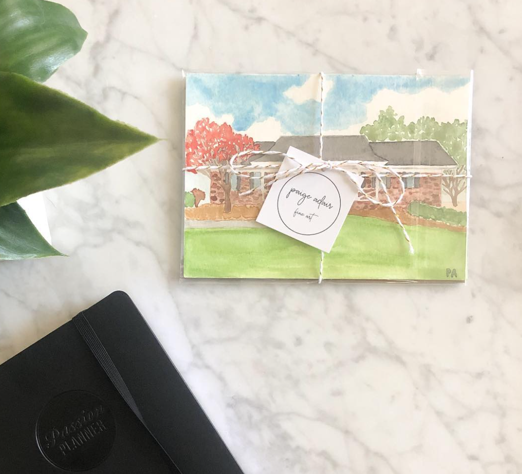 A wrapped art package with a watercolor house scene on the wrapping paper, tied with white and brown string, and a small white tag with handwritten text. A black dotted journal and a green plant leaf are on a marble surface.