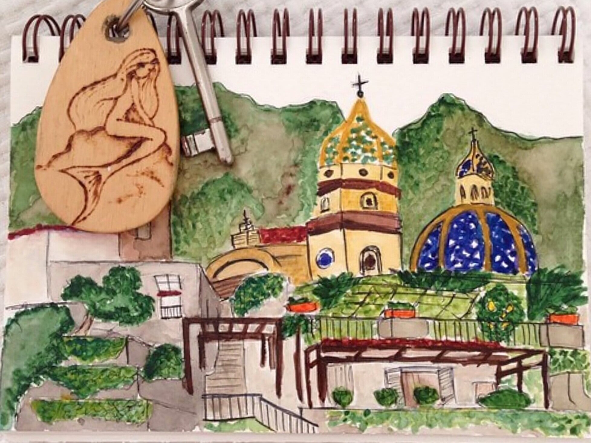 A colorful watercolor painting of a hillside with buildings, including two church domes and towers, surrounded by greenery, with a wooden owl-shaped pin and a silver pen placed on top of the painting.