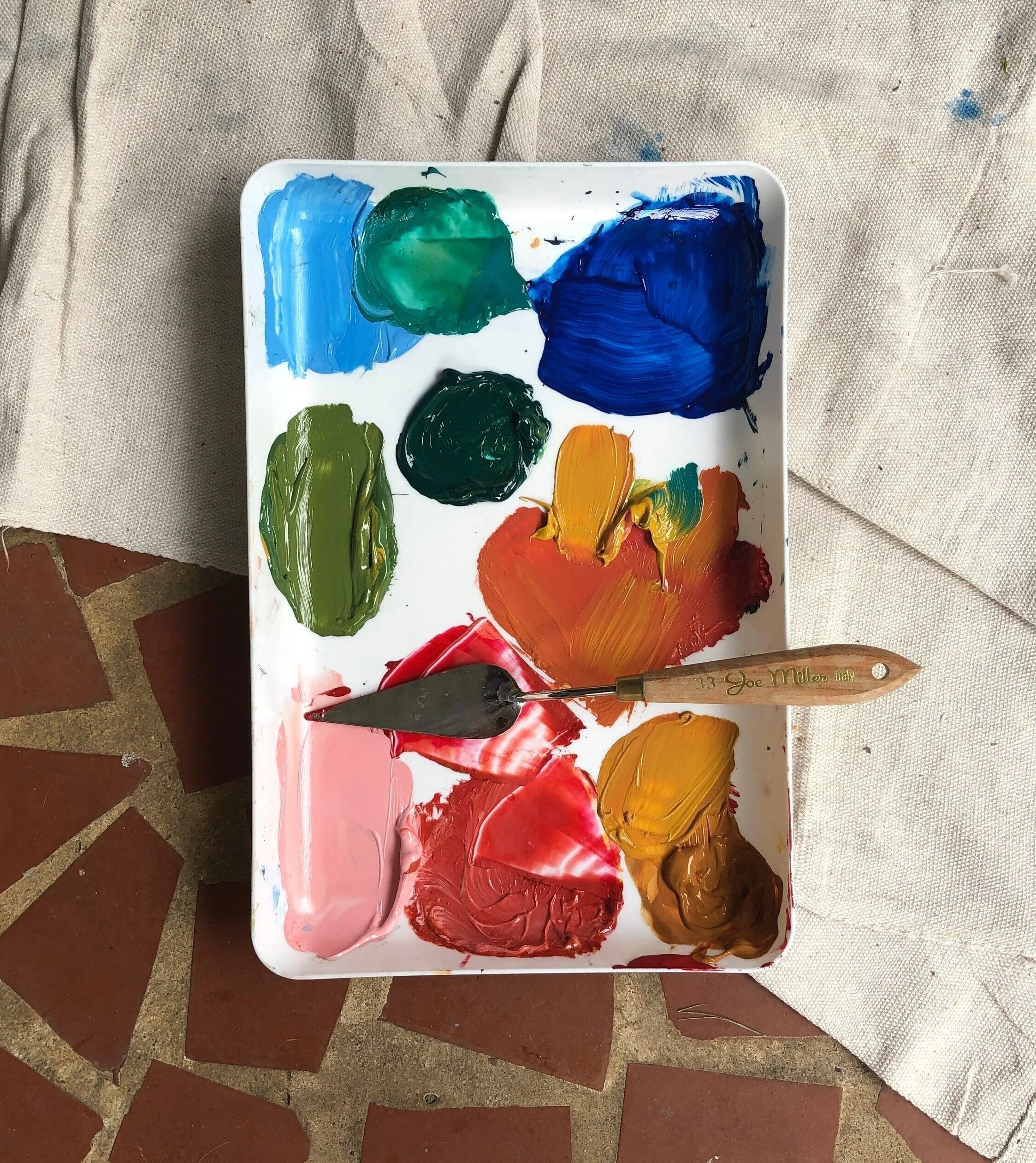 A white paint palette with various blobs of colorful paints, including blue, green, yellow, orange, red, brown, and pink, with a palette knife resting on it. The palette is on a tiled floor next to a piece of beige canvas.