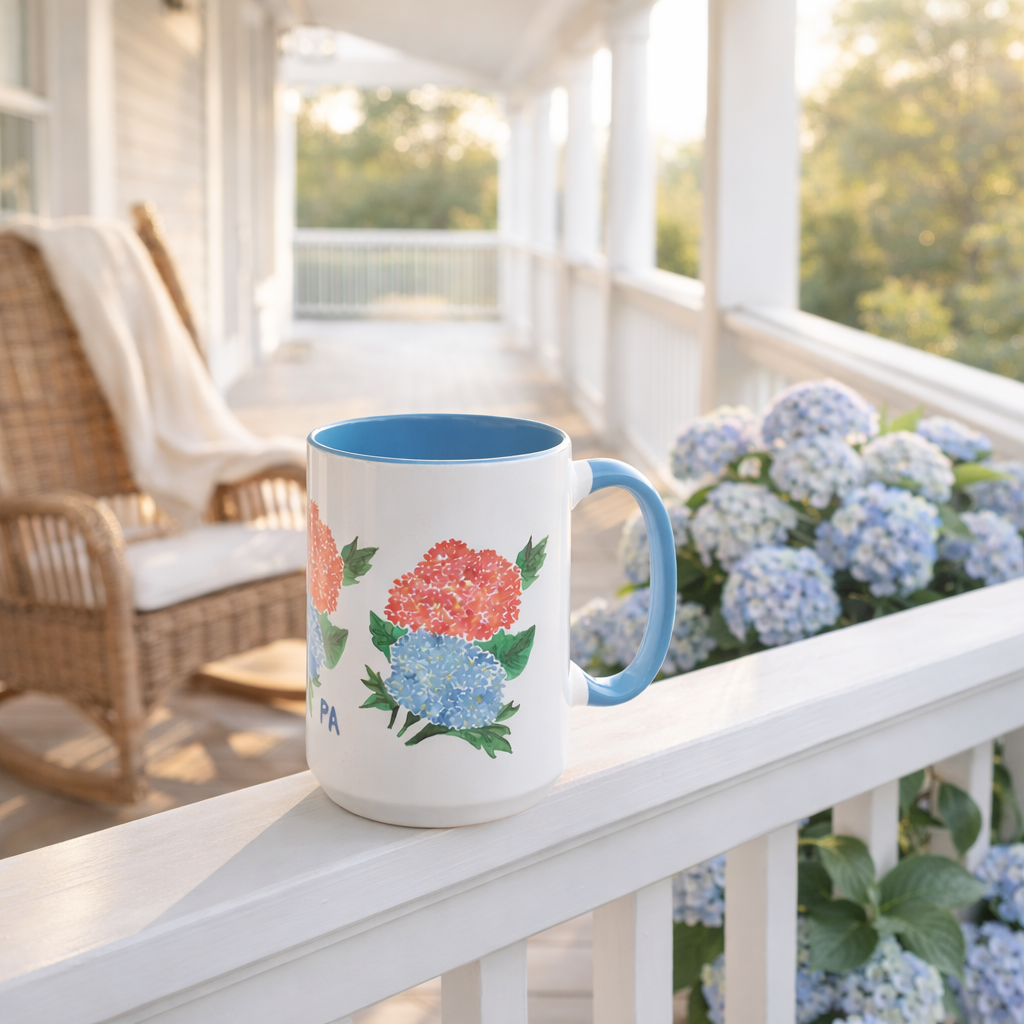 A ceramic mug with a floral design of pink and blue hydrangeas on a porch railing, with a background of white hydrangea flowers and a wicker chair on a porch.