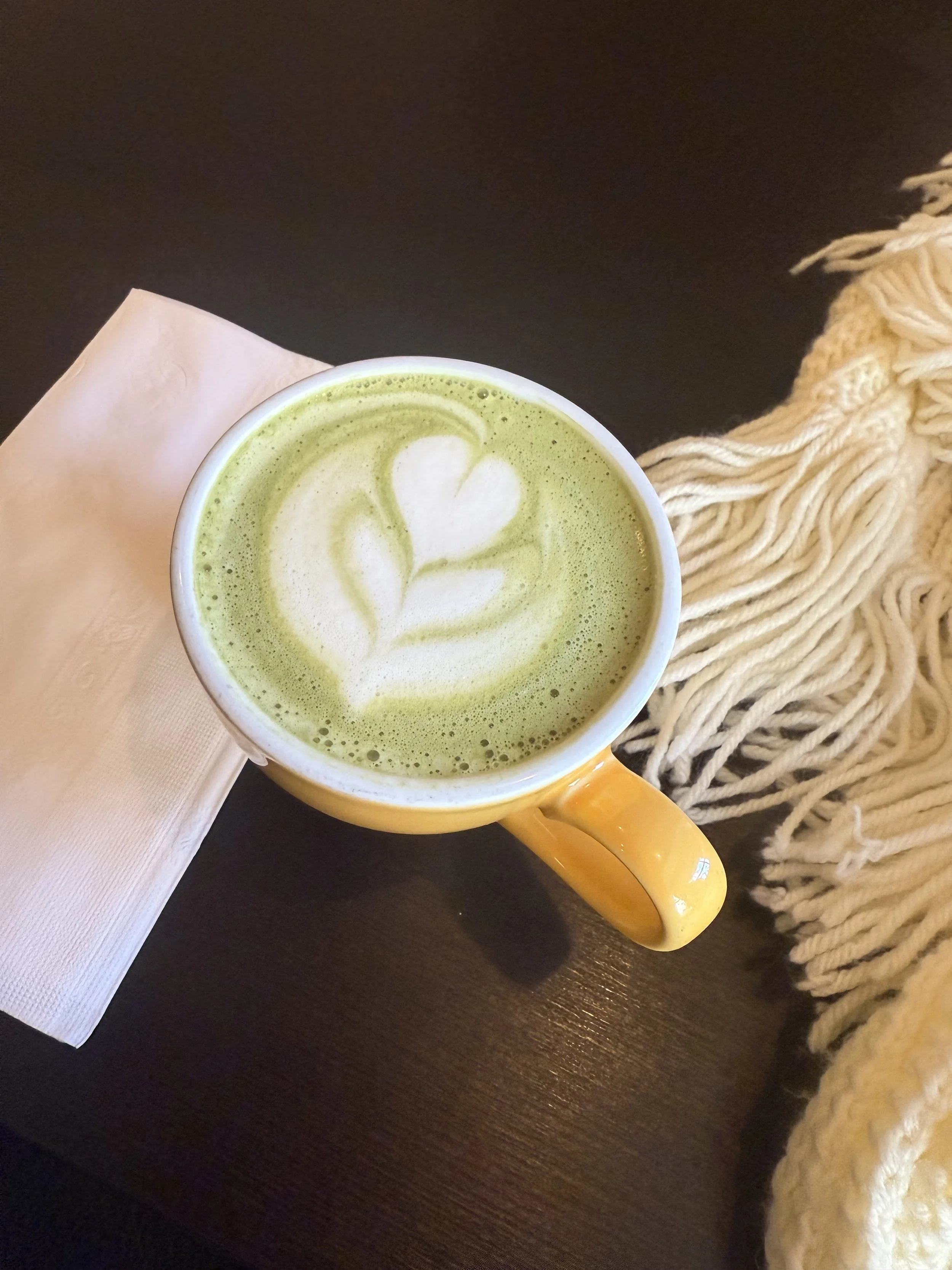 A hot matcha latte in a yellow mug with steamed milk art in the shape of a heart. A napkin and scarf to the sides.