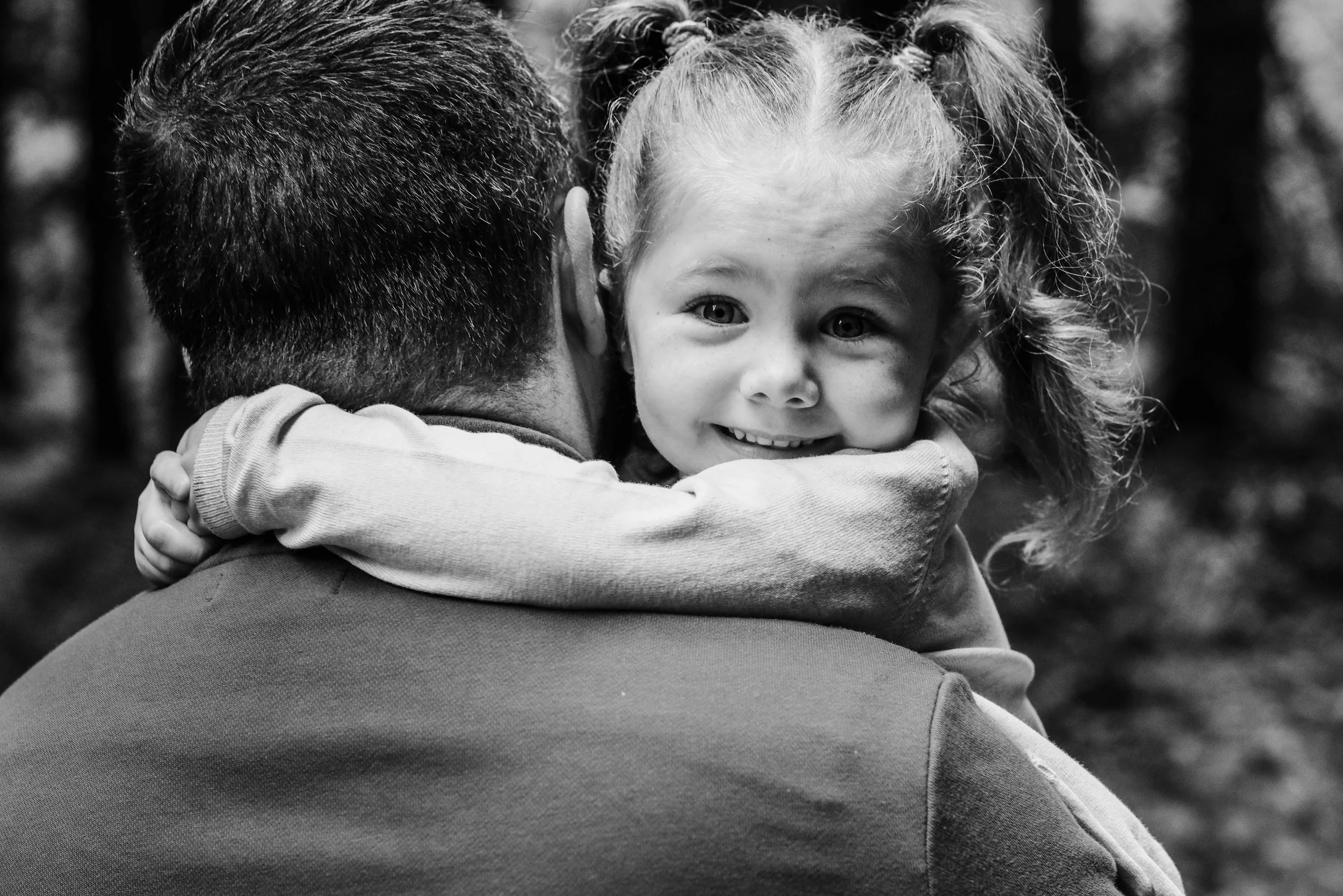 A man holding a smiling young girl in an outdoor setting, with trees in the background.