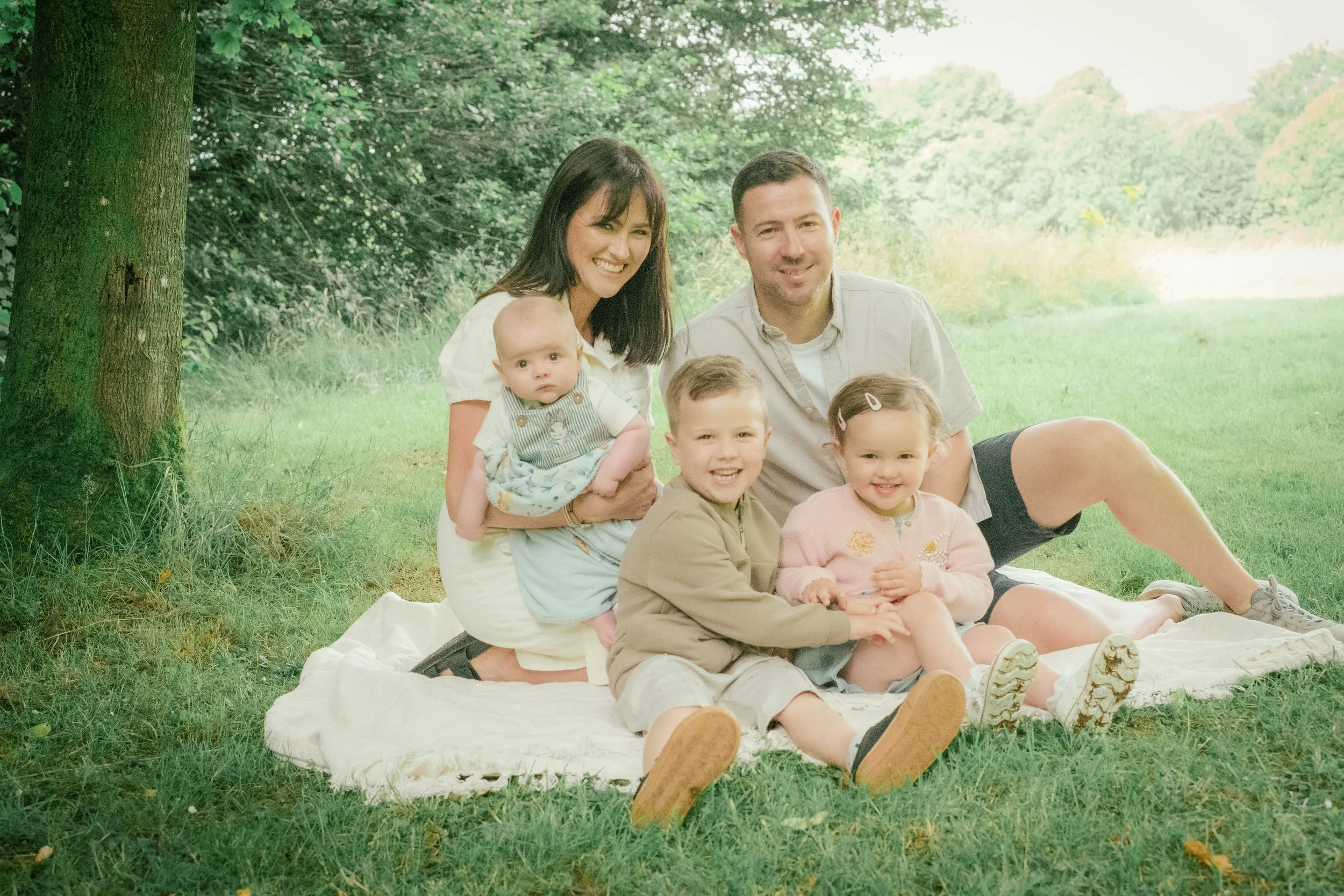 A family of two adults and three children sitting on a white blanket outdoors near a tree, with green grass and trees in the background, on a sunny day.