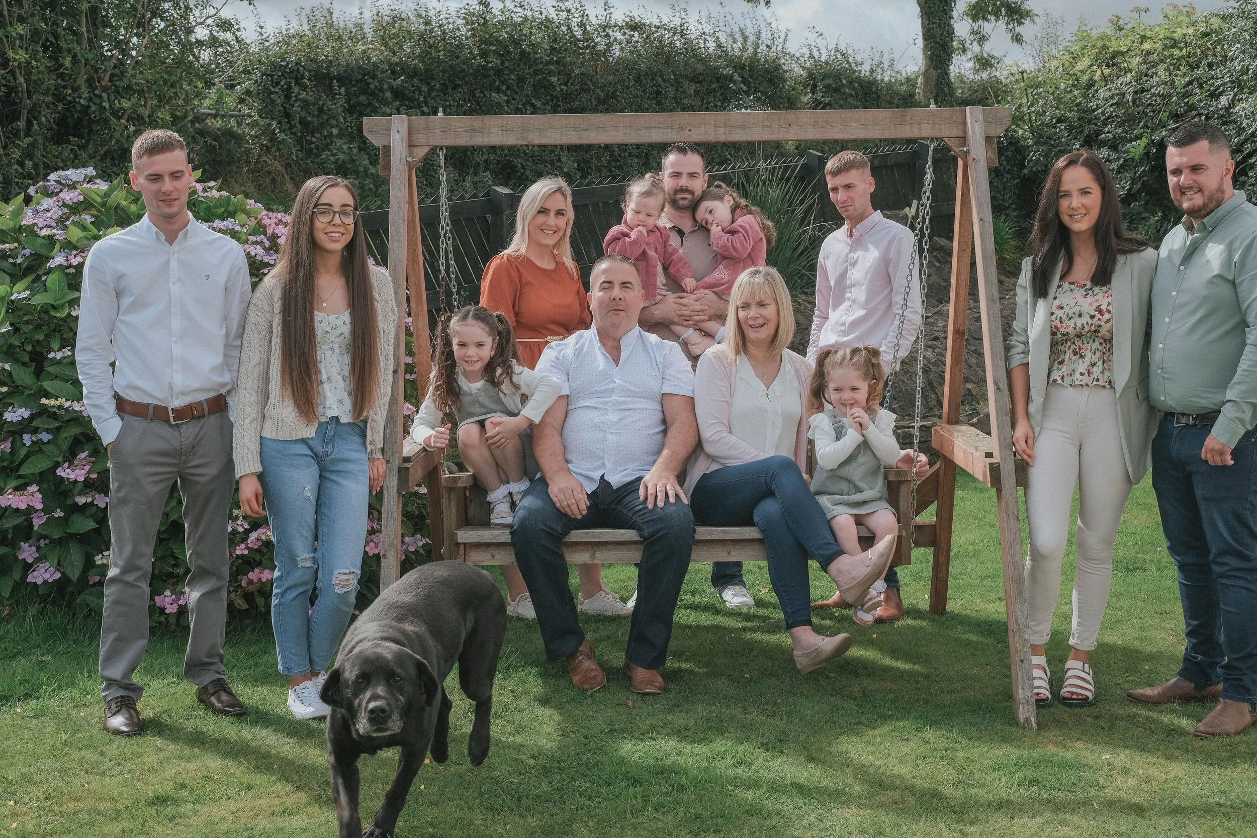 A large multi-generational family gathered outdoors on a cloudy day, with some members standing and others sitting on a wooden swing set, surrounded by greenery and blooming hydrangeas, along with a black Labrador retriever in the foreground.