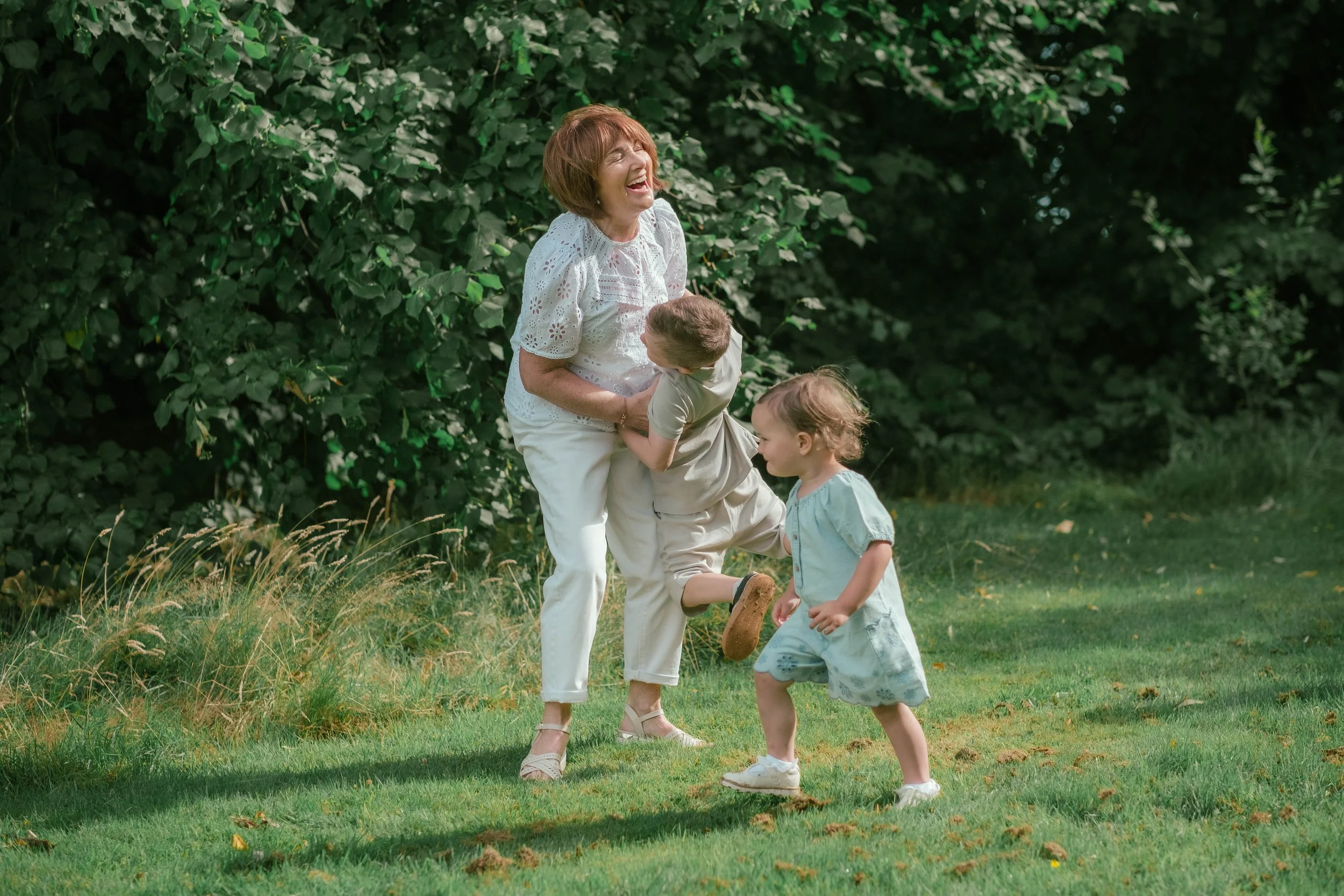 A woman and two children laughing and playing outside on a grassy area with green bushes in the background.
