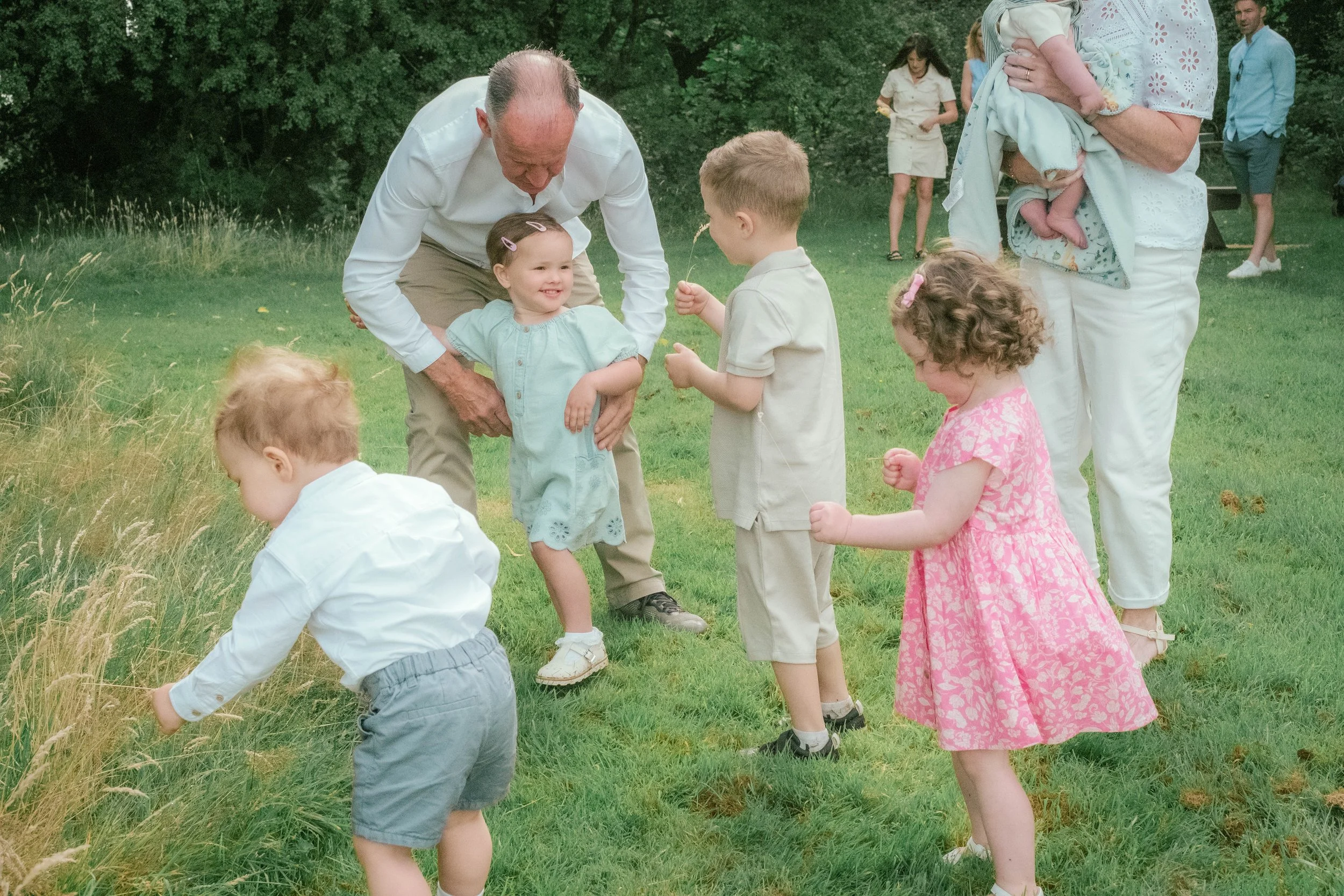 A group of children and adults outdoors on a grassy field, engaging with plants or grass, with trees in the background.