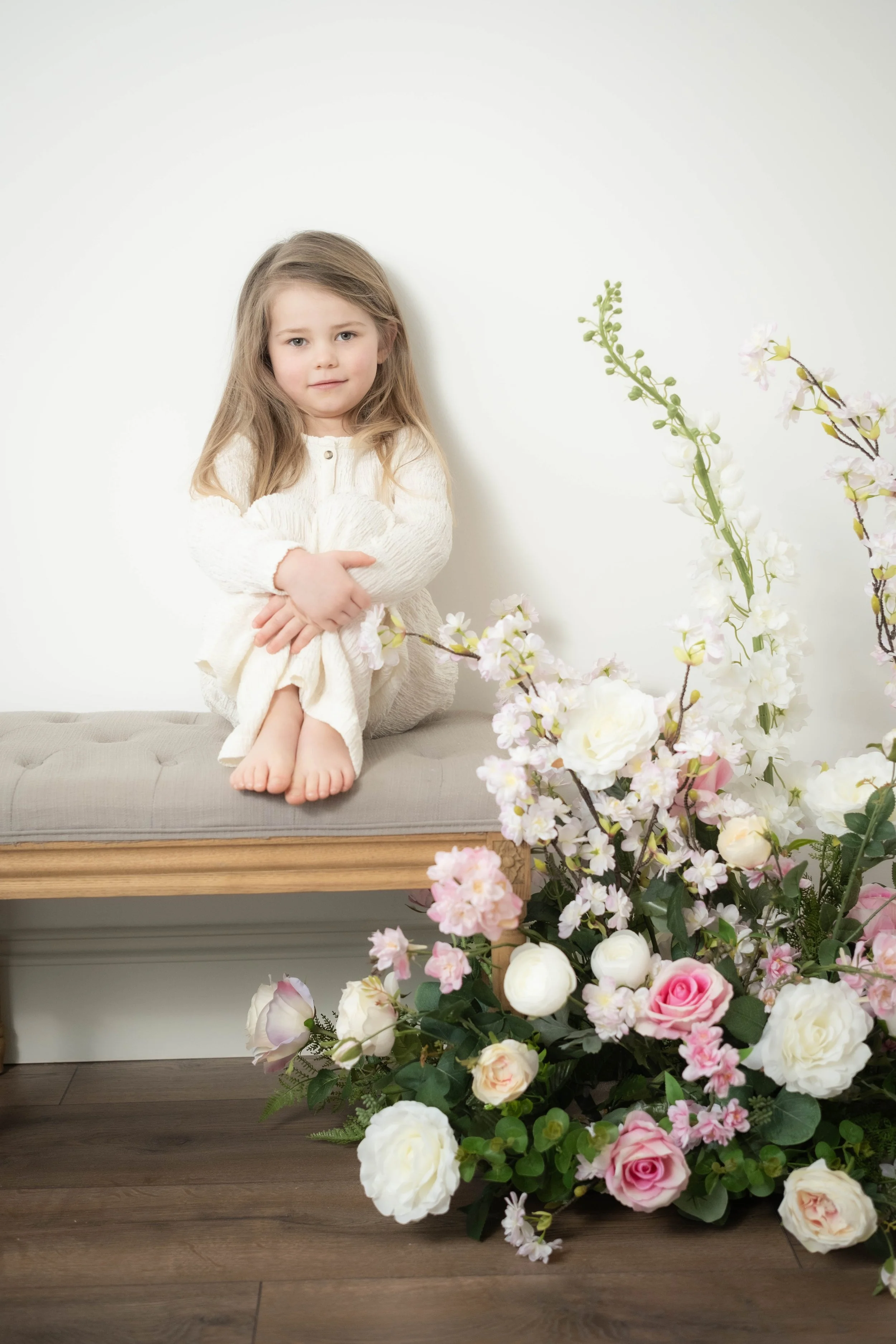 A young girl sitting on a beige bench with crossed arms, next to a large bouquet of pink and white flowers, against a plain white wall.
