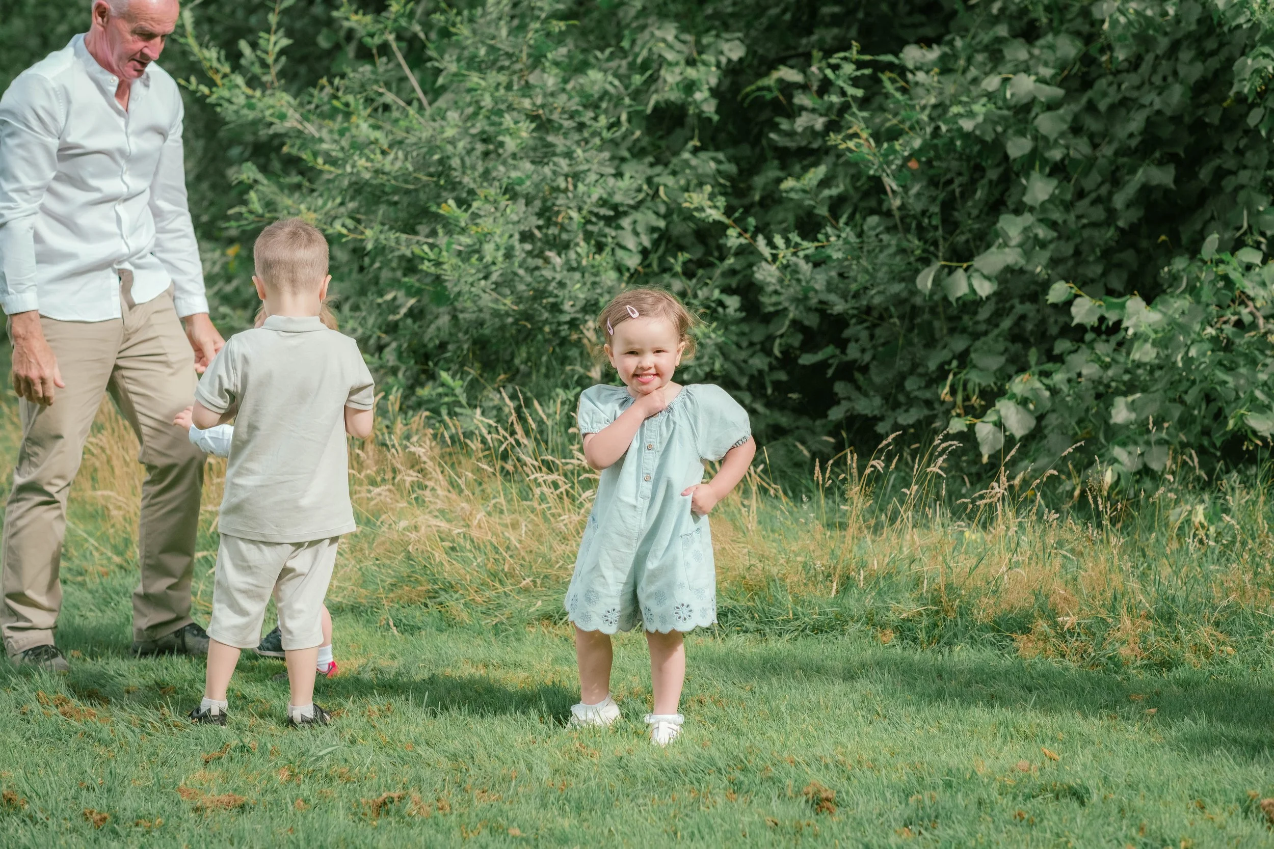 A young girl smiling and standing with one hand on her chin outdoors, with a man and two boys in the background near green shrubbery and grass.