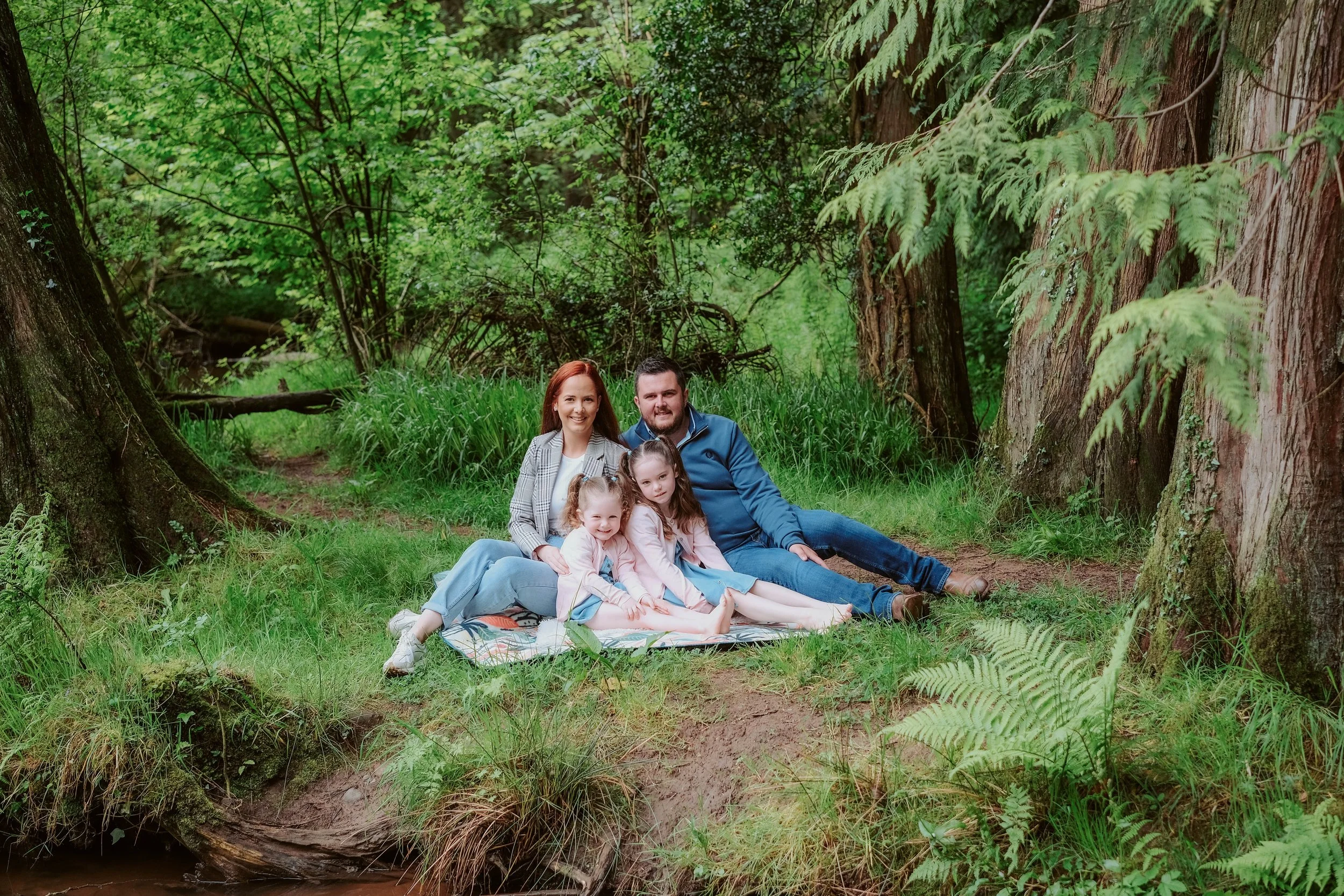 Family of four sitting on a blanket in a lush green forest.