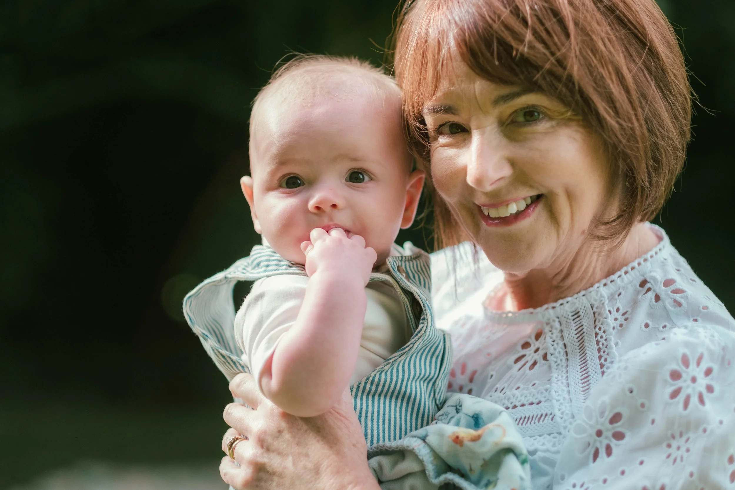 An woman with short, reddish hair smiling while holding a baby with light skin and big eyes, who is sucking on their hand, outdoors with a dark, blurred background.