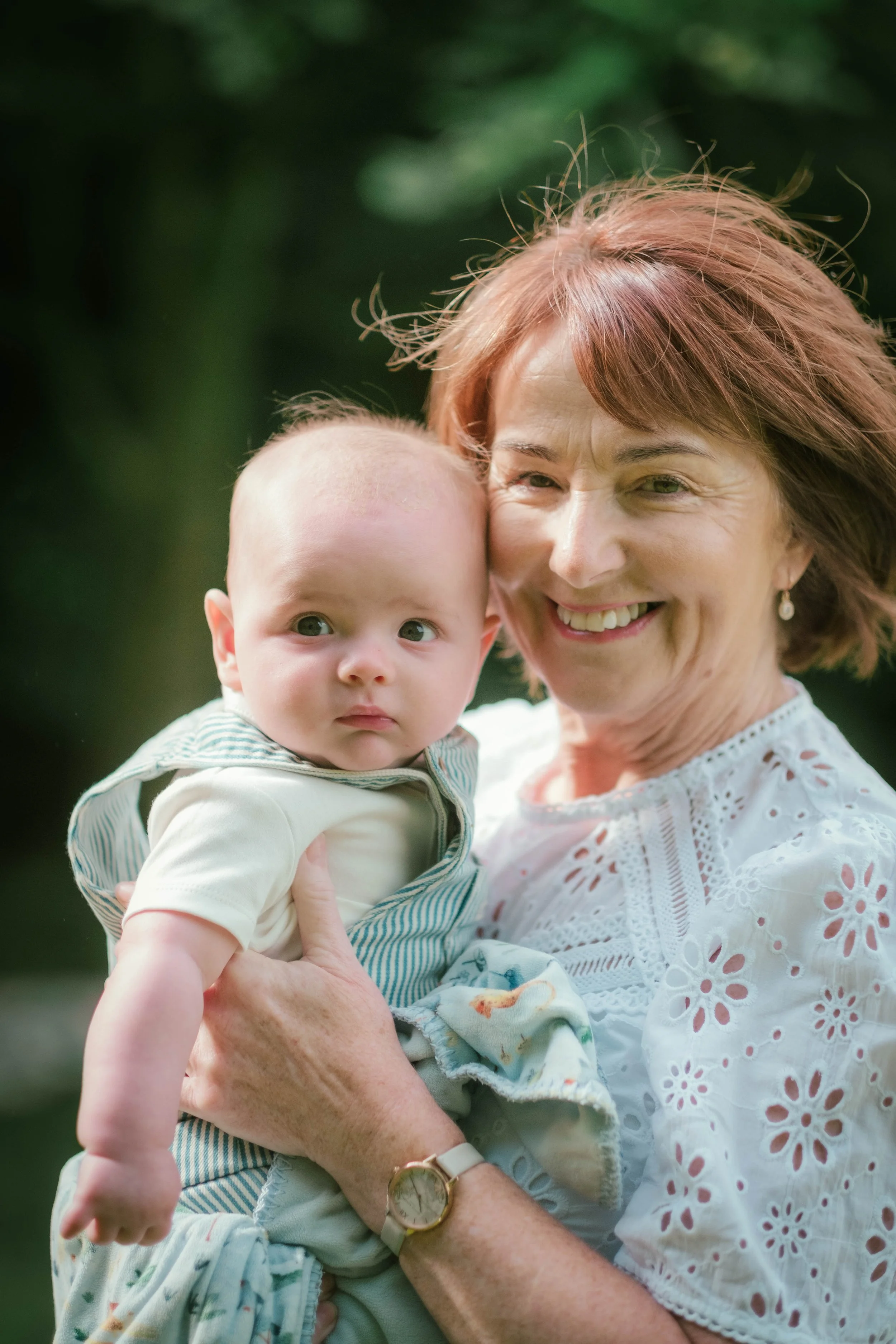 An older woman holding a baby outdoors with a blurred green background, both smiling and looking at the camera.