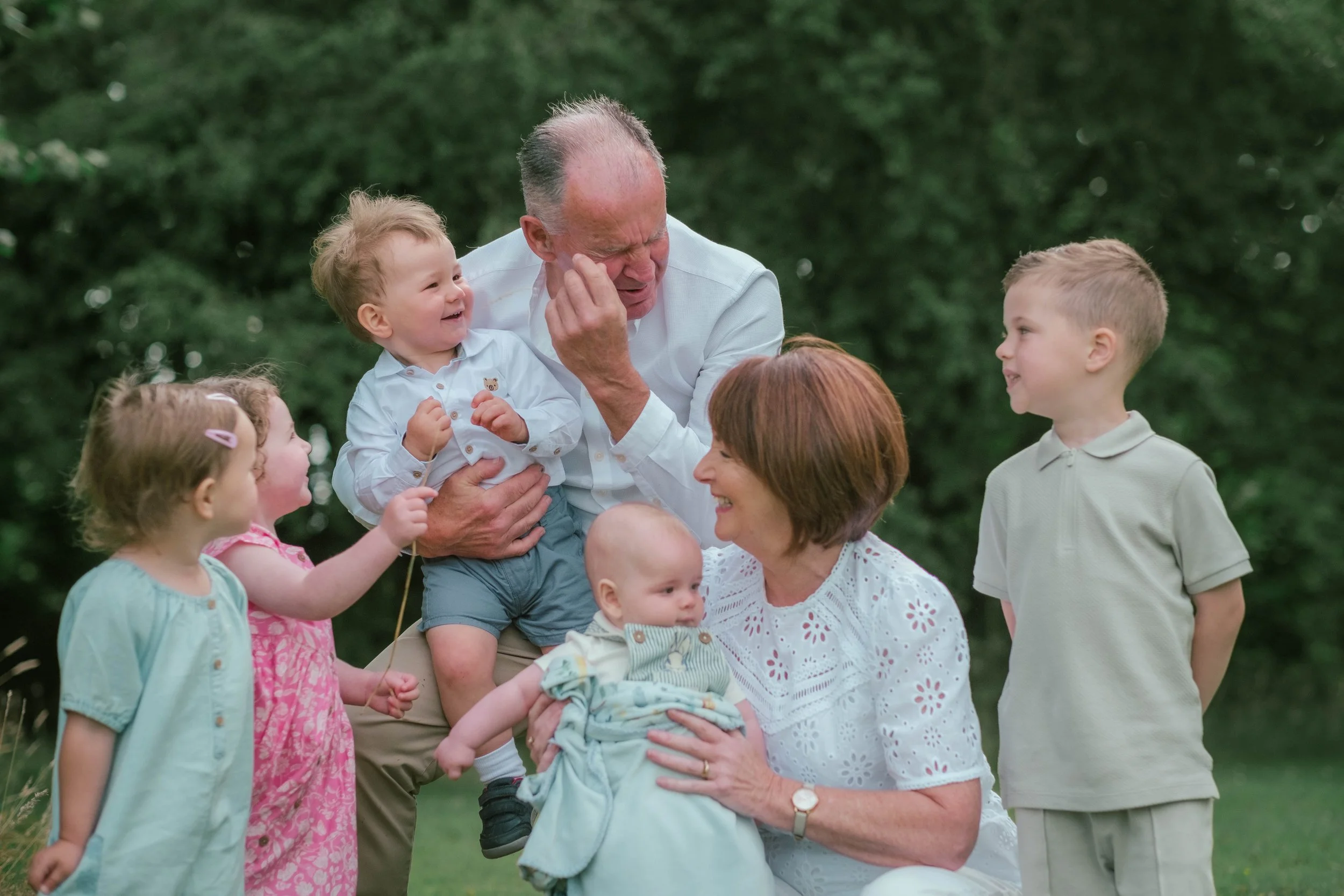 A multigenerational family outdoors, with grandparents and children, smiling and playing together on a grassy area with trees in the background.